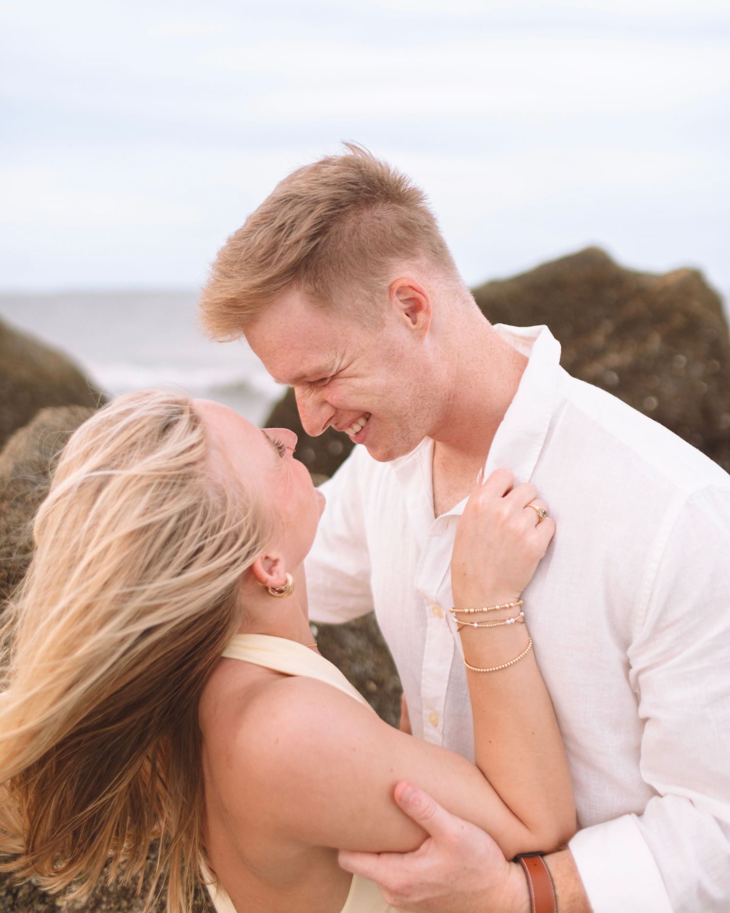 Smiling blonde couple embracing near rocky beach, woman in gold dress, man in white shirt, ocean backdrop.