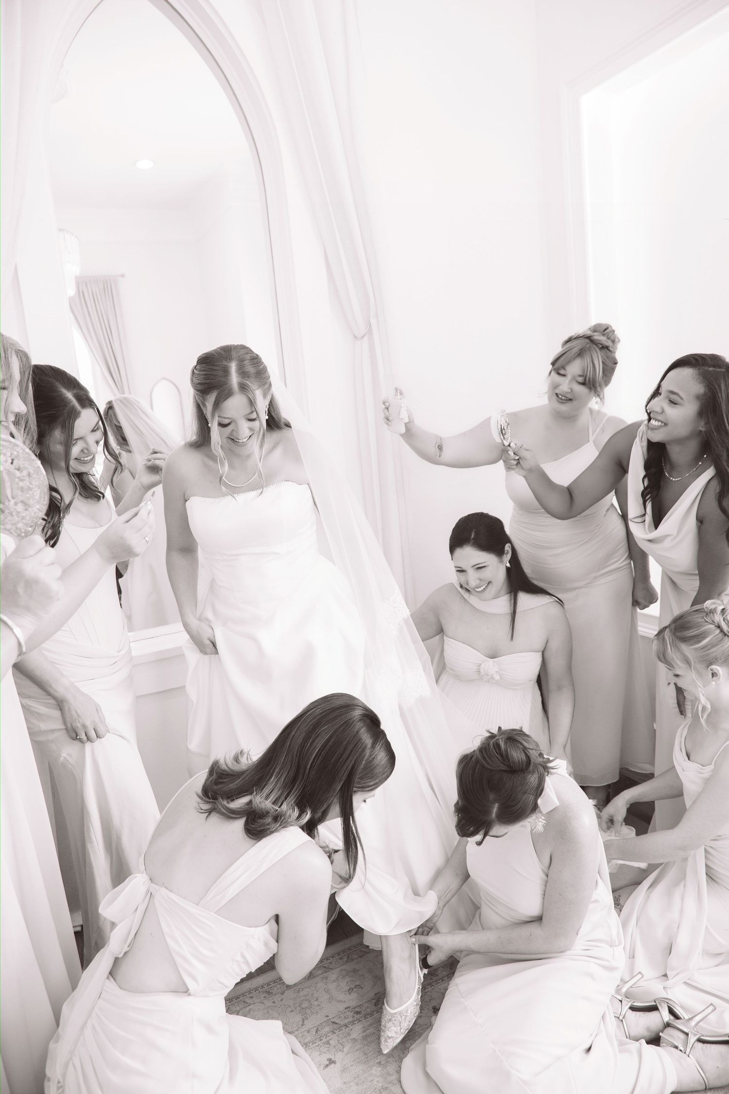 Bridesmaids help bride get ready in a bright white dressing room in this black and white wedding photo.