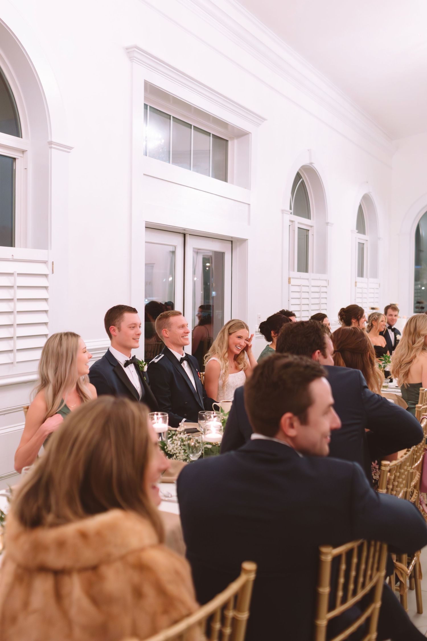 Wedding guests in formal attire seated at elegant reception tables in white arched venue.