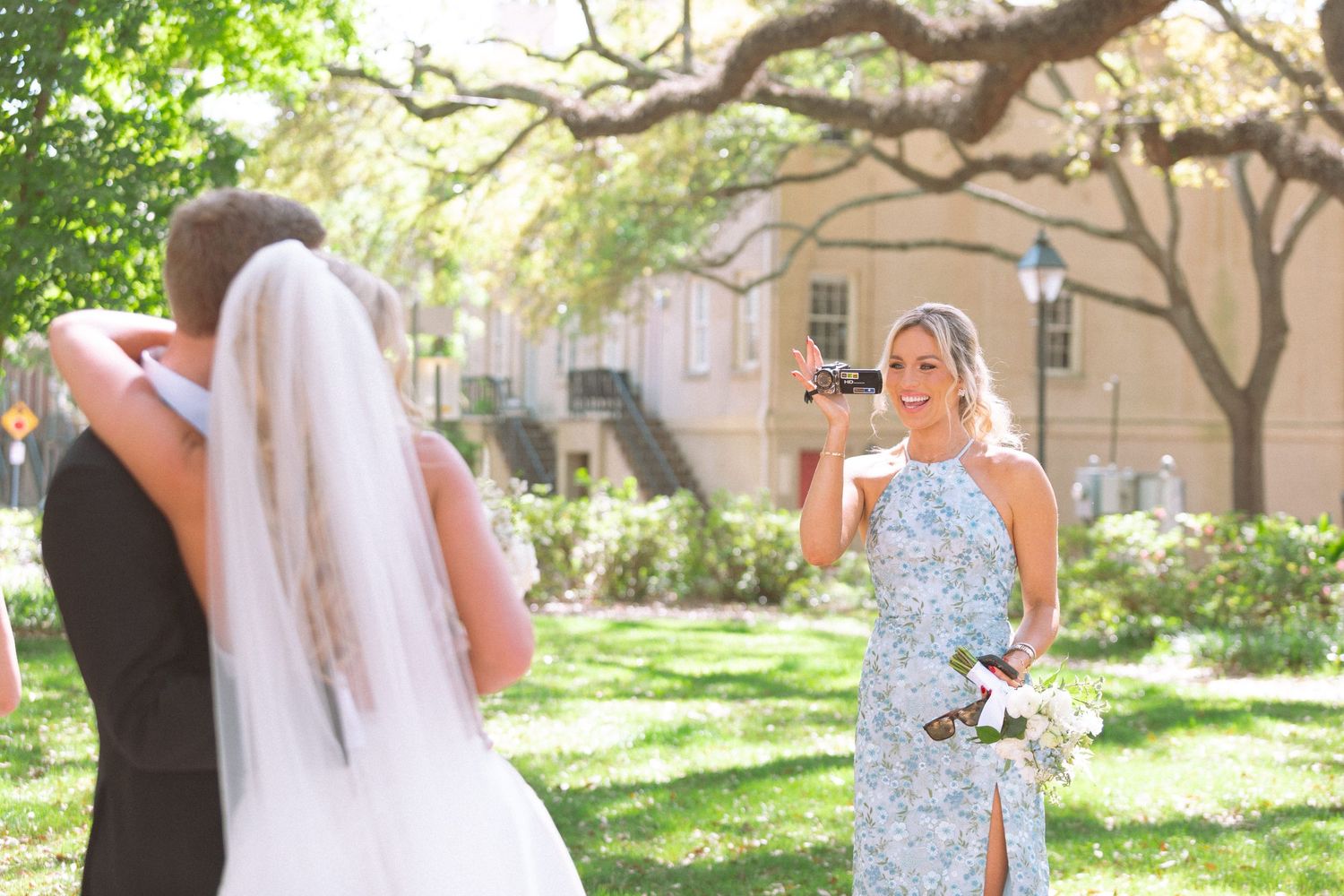 Woman in floral dress photographs bride and groom embracing outdoors at a sunny wedding ceremony.