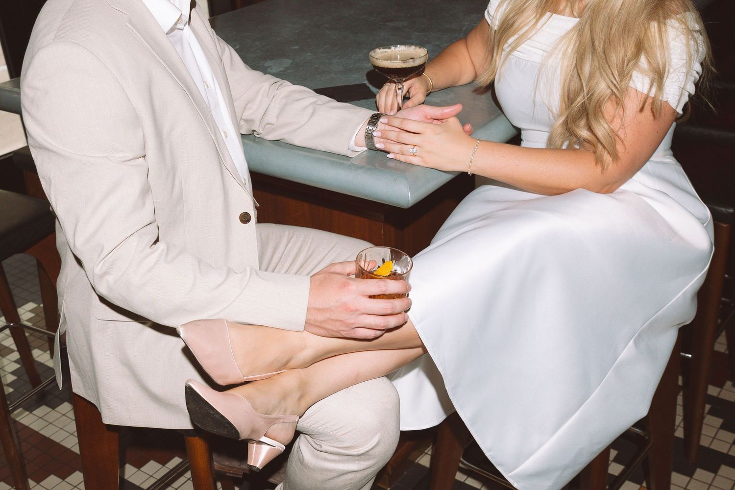 Bride and groom sitting at a bar, holding drinks, dressed in white wedding attire and beige suit.