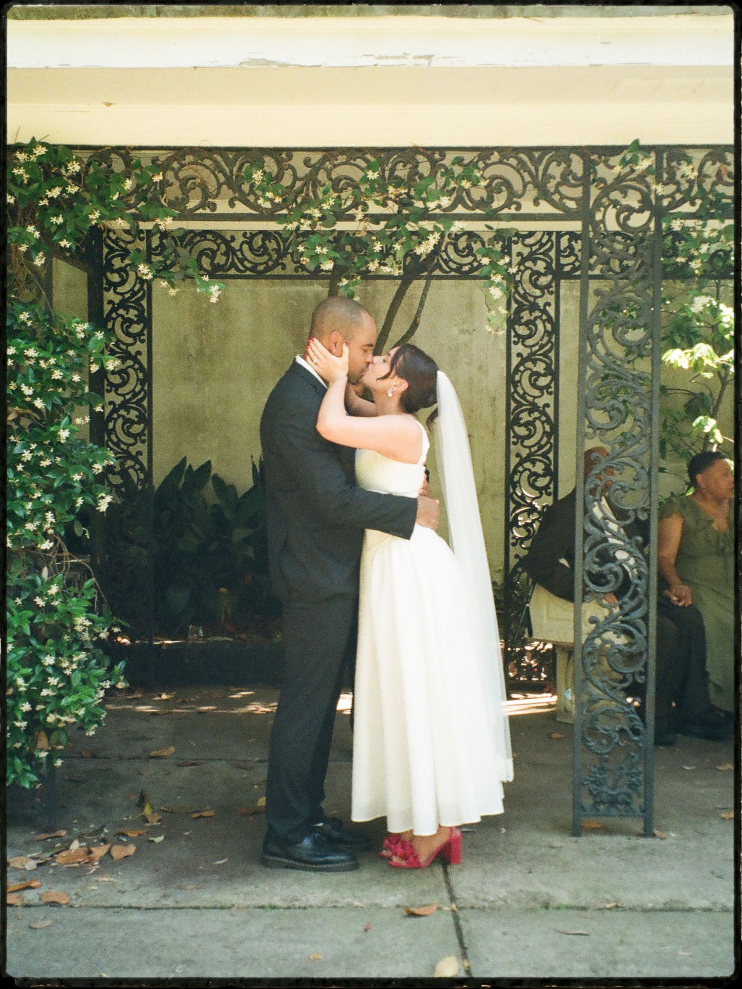 Newlyweds share first kiss beneath ornate wrought iron gate with lush ivy backdrop.