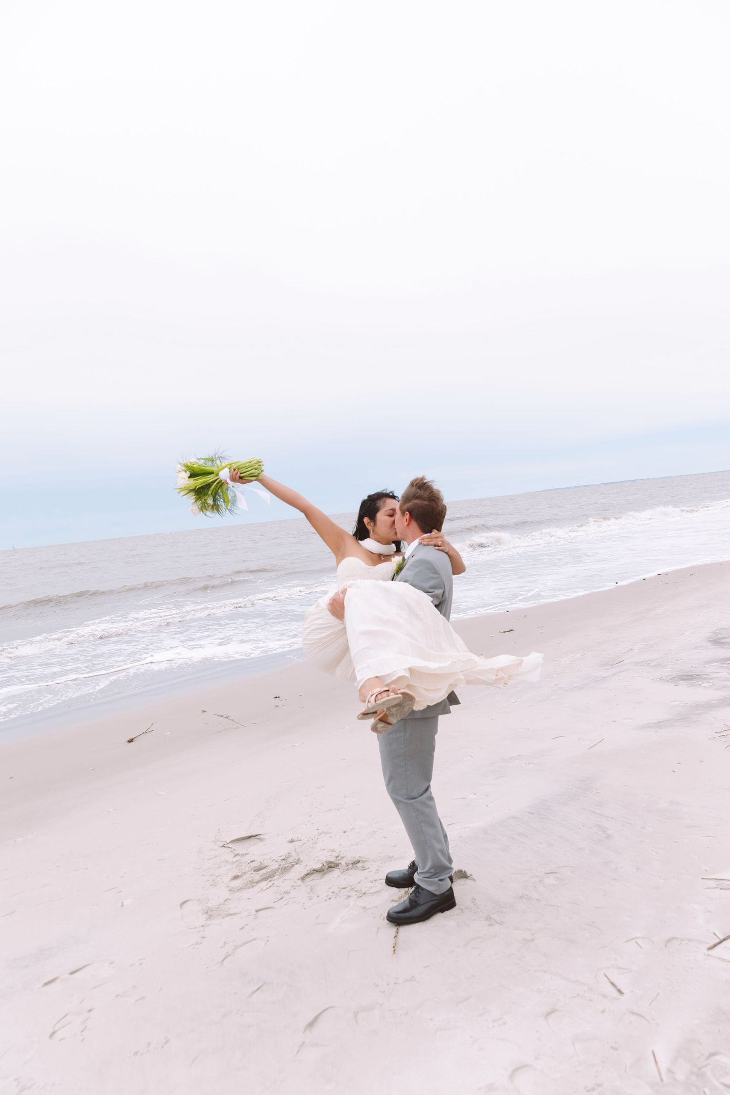 Groom lifting bride on a sandy beach as she raises her bouquet, sharing a romantic kiss by the ocean.