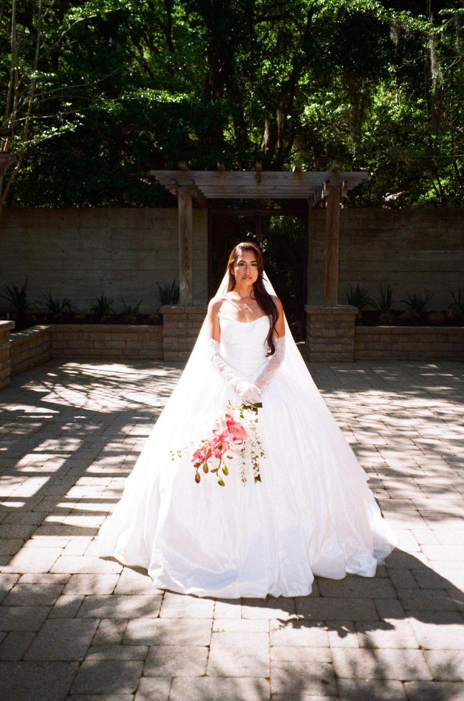 Bride in a white ball gown holding a pink bouquet stands on a stone patio before a wooden arbor surrounded by lush trees.