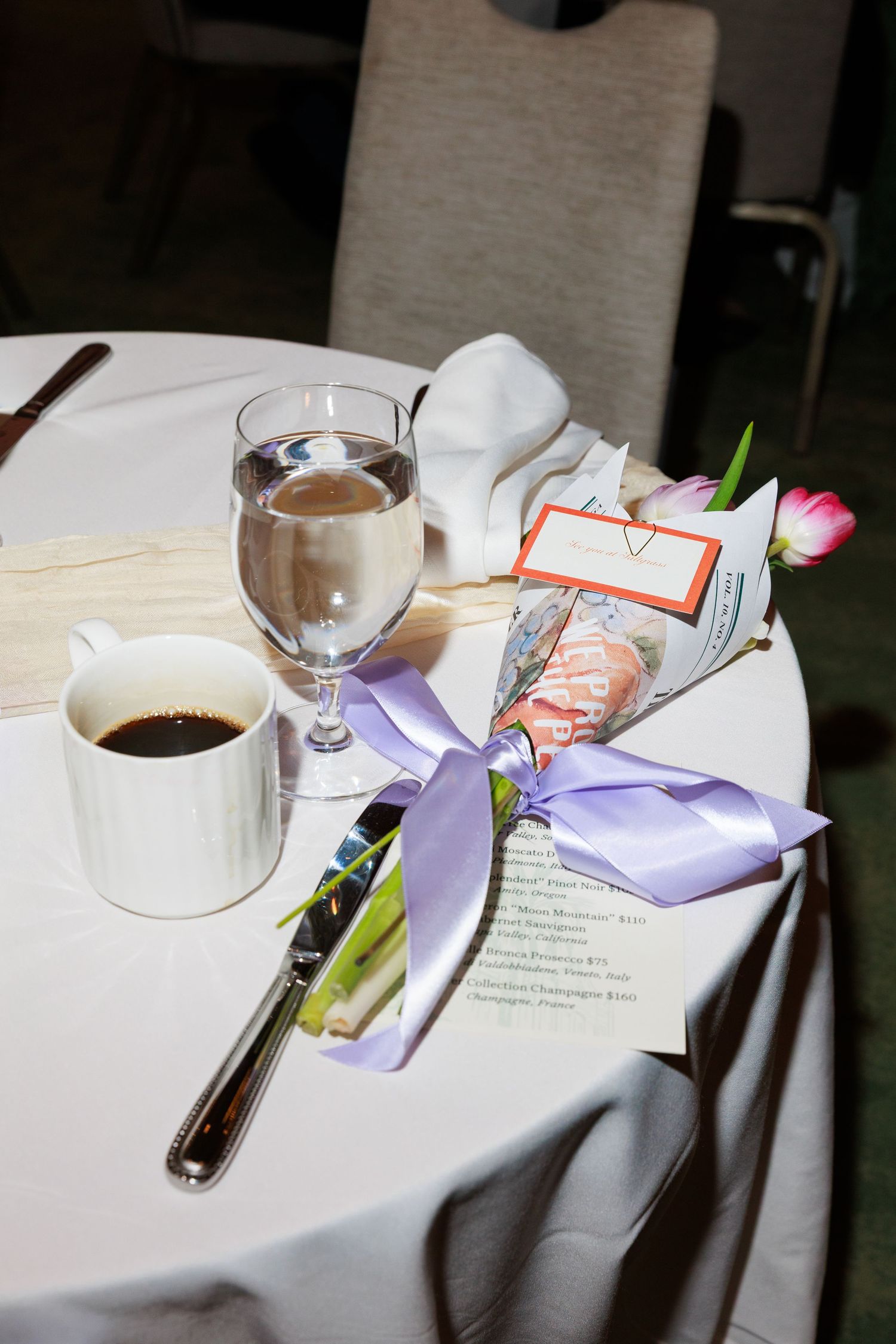 Elegant table setting with coffee, water glass, tulip bouquet tied with purple ribbon, and menu card at a formal event.