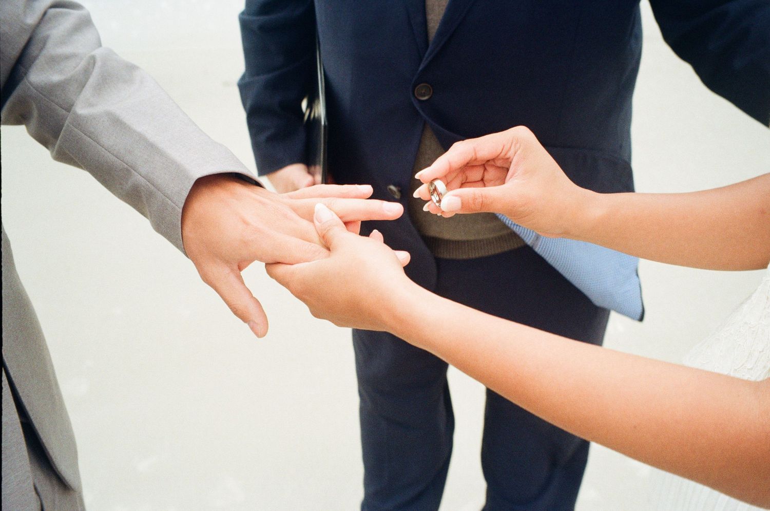 A bride placing a wedding ring on the groom's finger during a ceremony, officiated by a person in a navy suit.