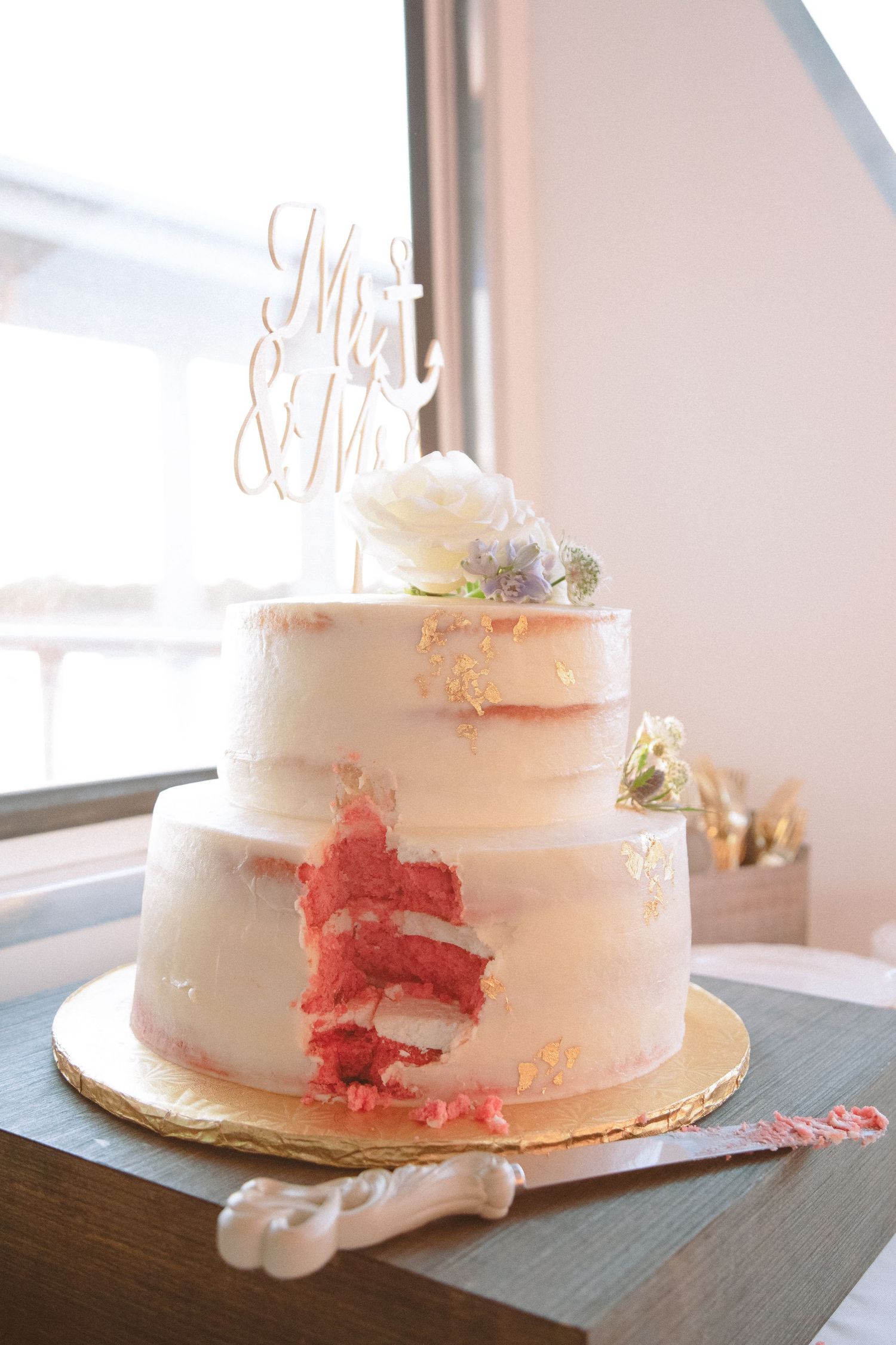 Elegant two-tier naked wedding cake with Mr and Mrs topper and floral decorations on wooden table.
