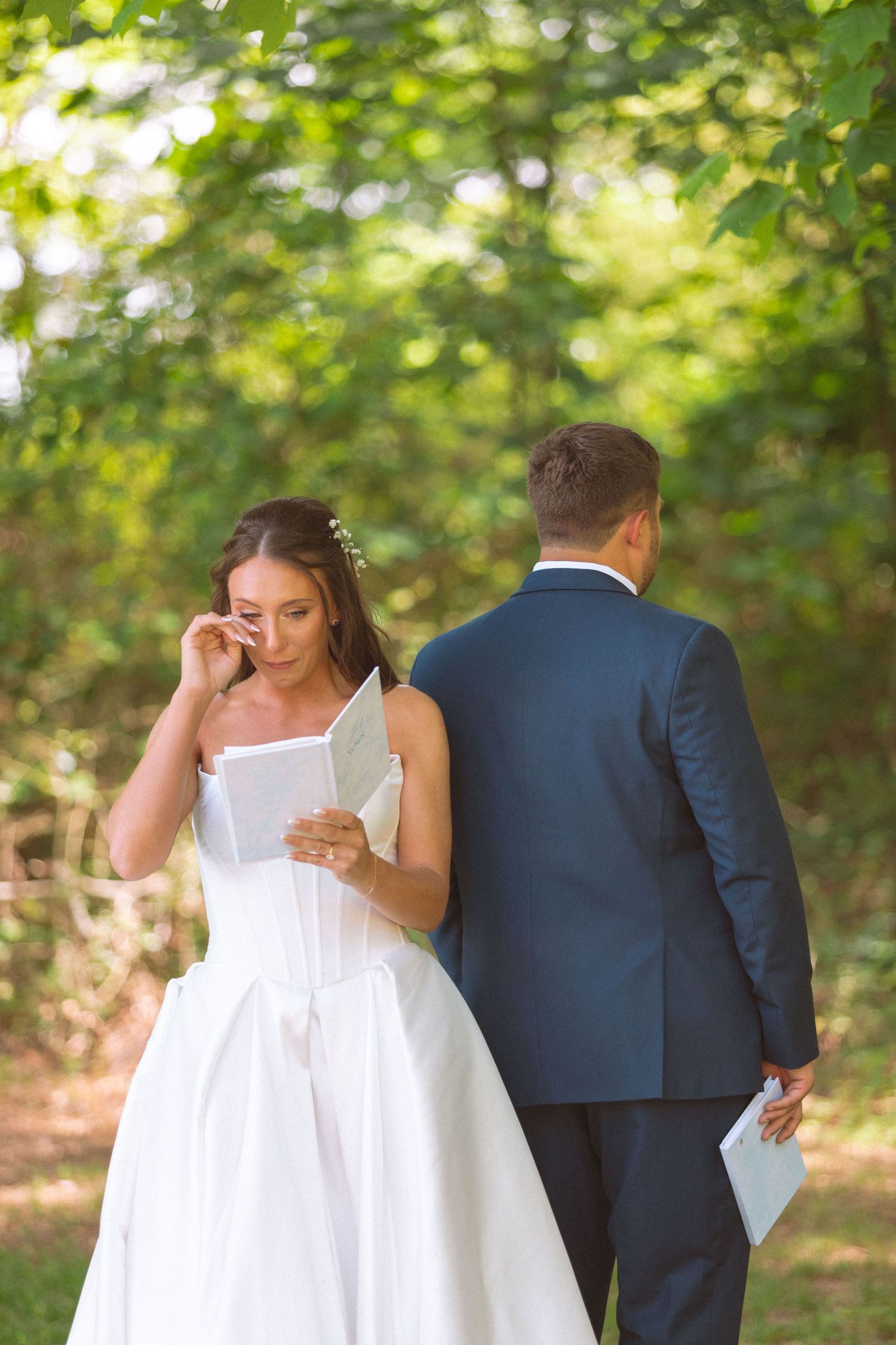 Emotional bride wipes tears reading vows while groom stands back-to-back in green garden setting.