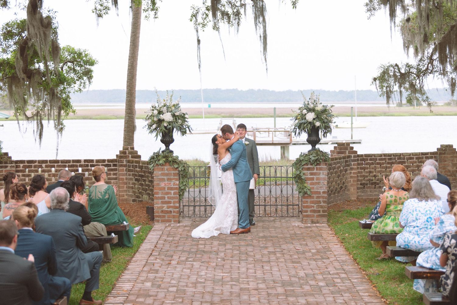 Bride and groom share first kiss at outdoor waterfront wedding ceremony with brick pathway and Spanish moss trees.