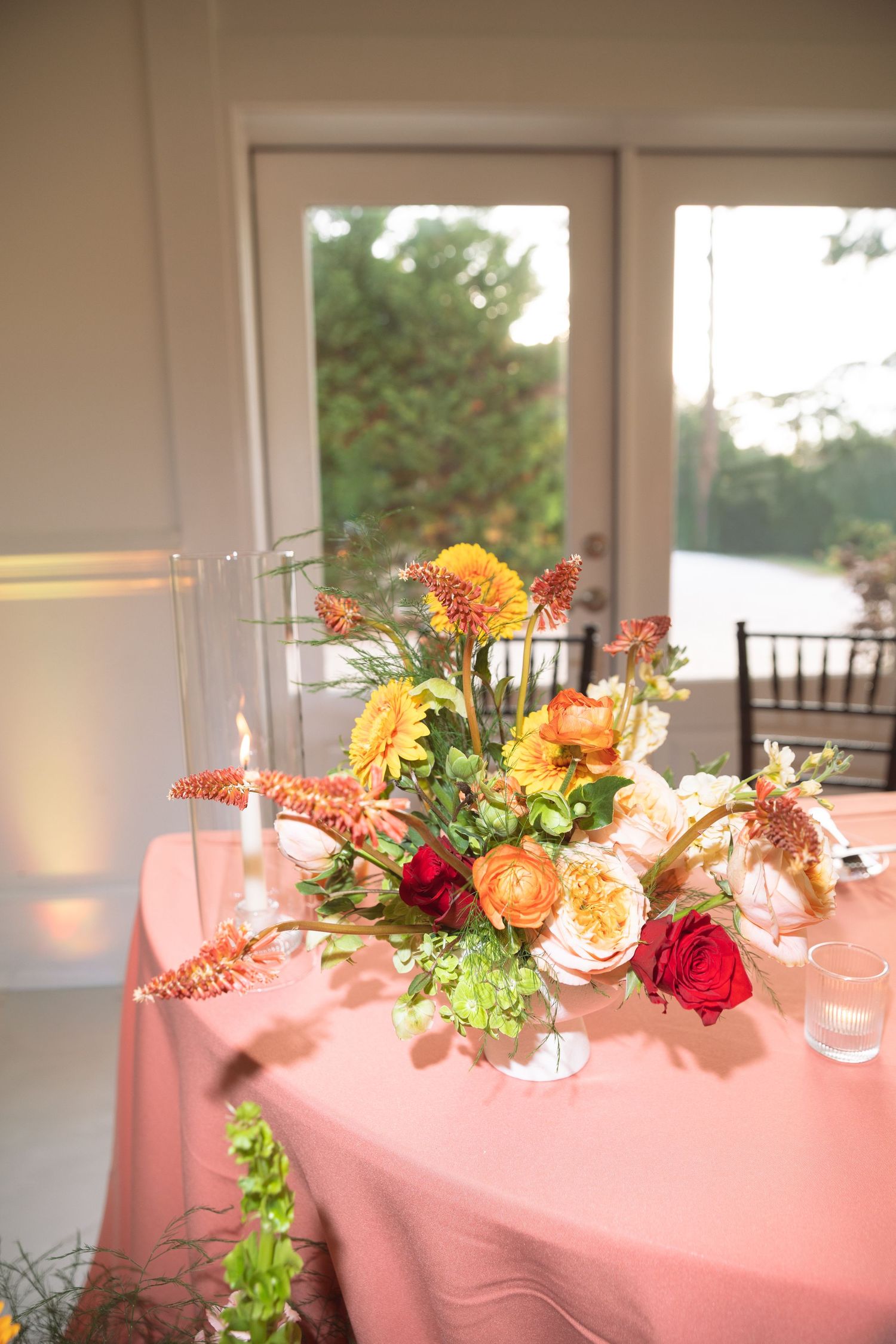 Vibrant wedding centerpiece featuring orange, red, and yellow flowers on a pink tablecloth near a window.