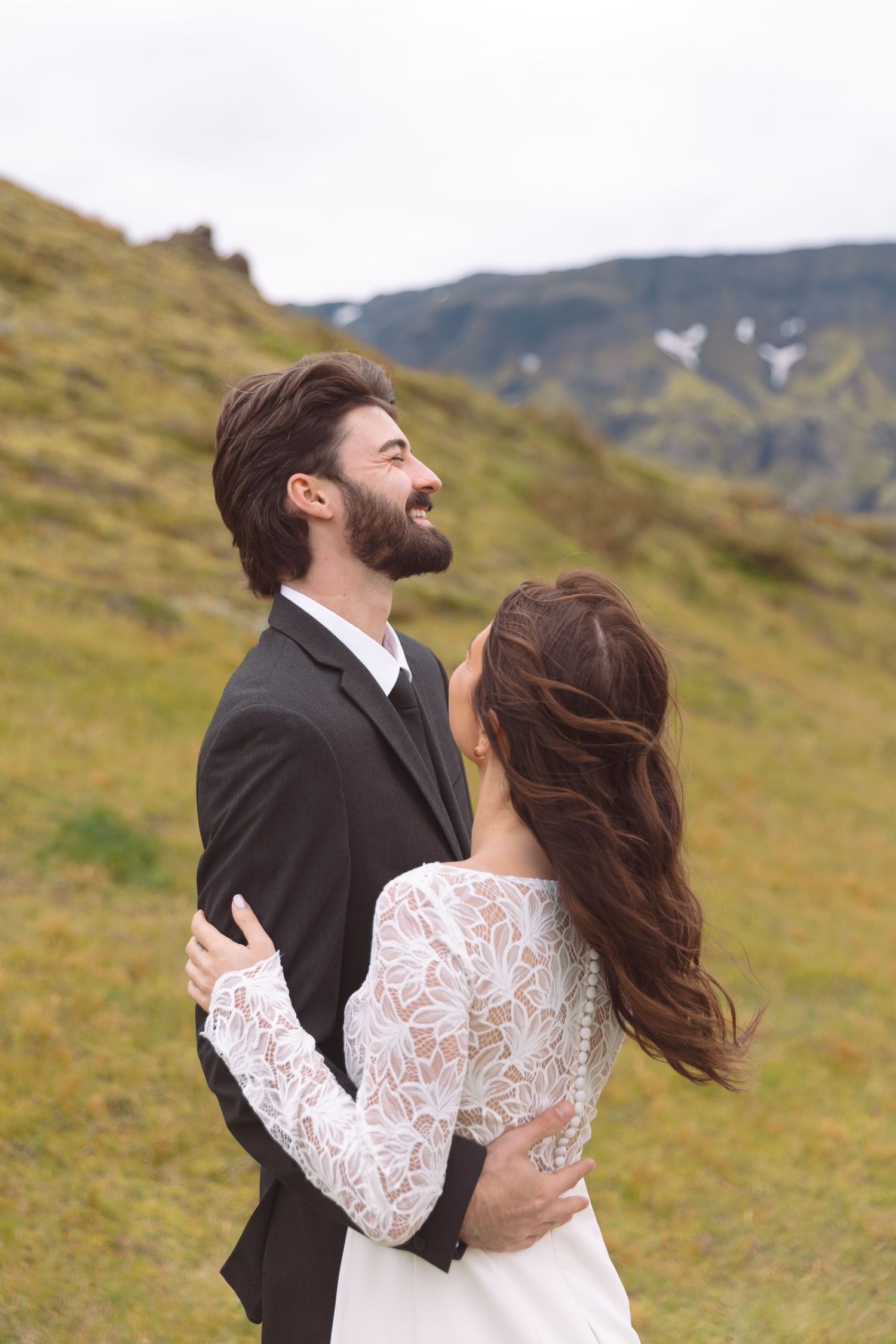 Laughing groom in dark suit embraces bride in lace wedding dress on scenic green hillside.