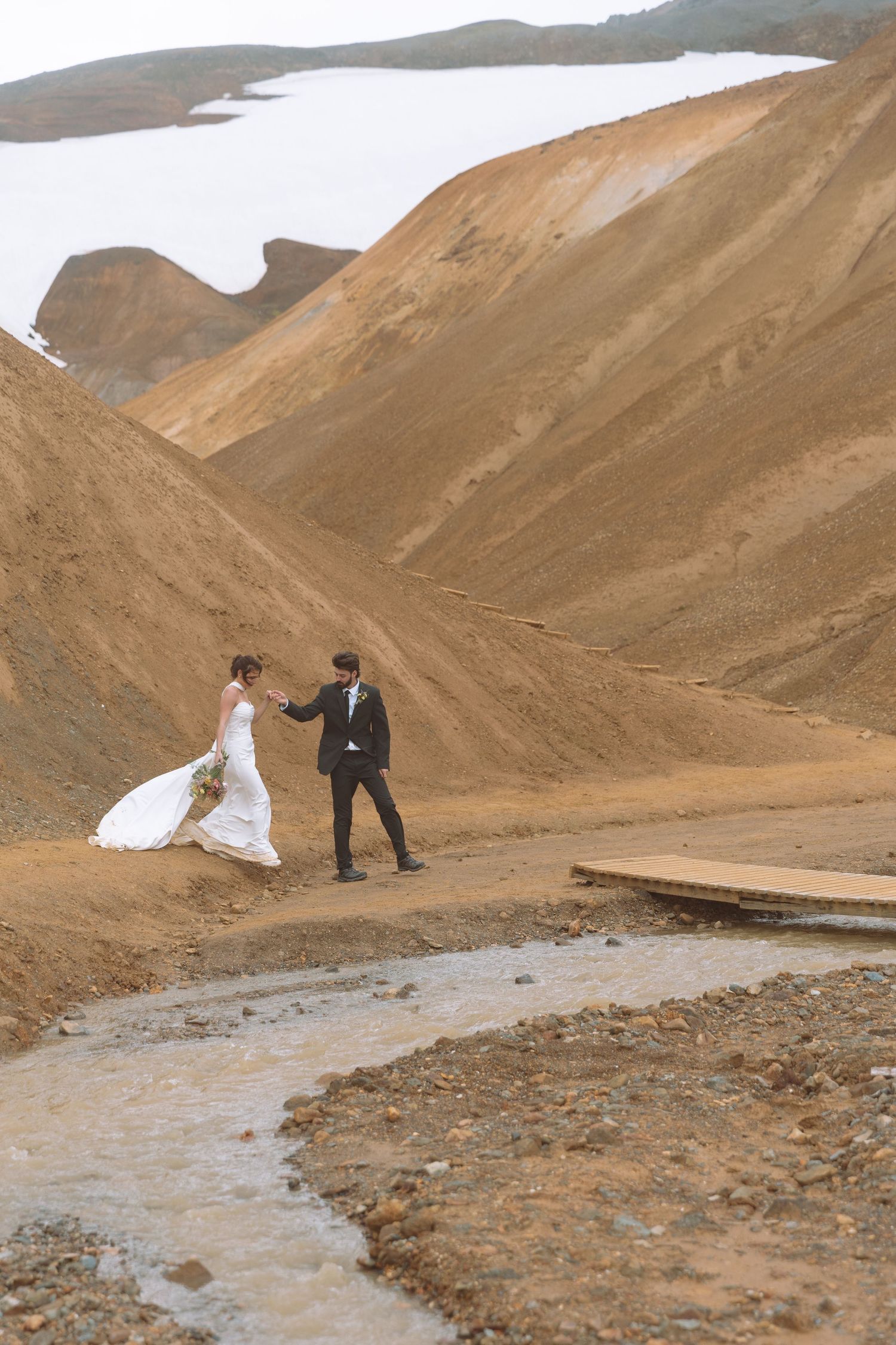 Bride and groom hold hands near a stream in Iceland's dramatic rhyolite mountains during their destination wedding.