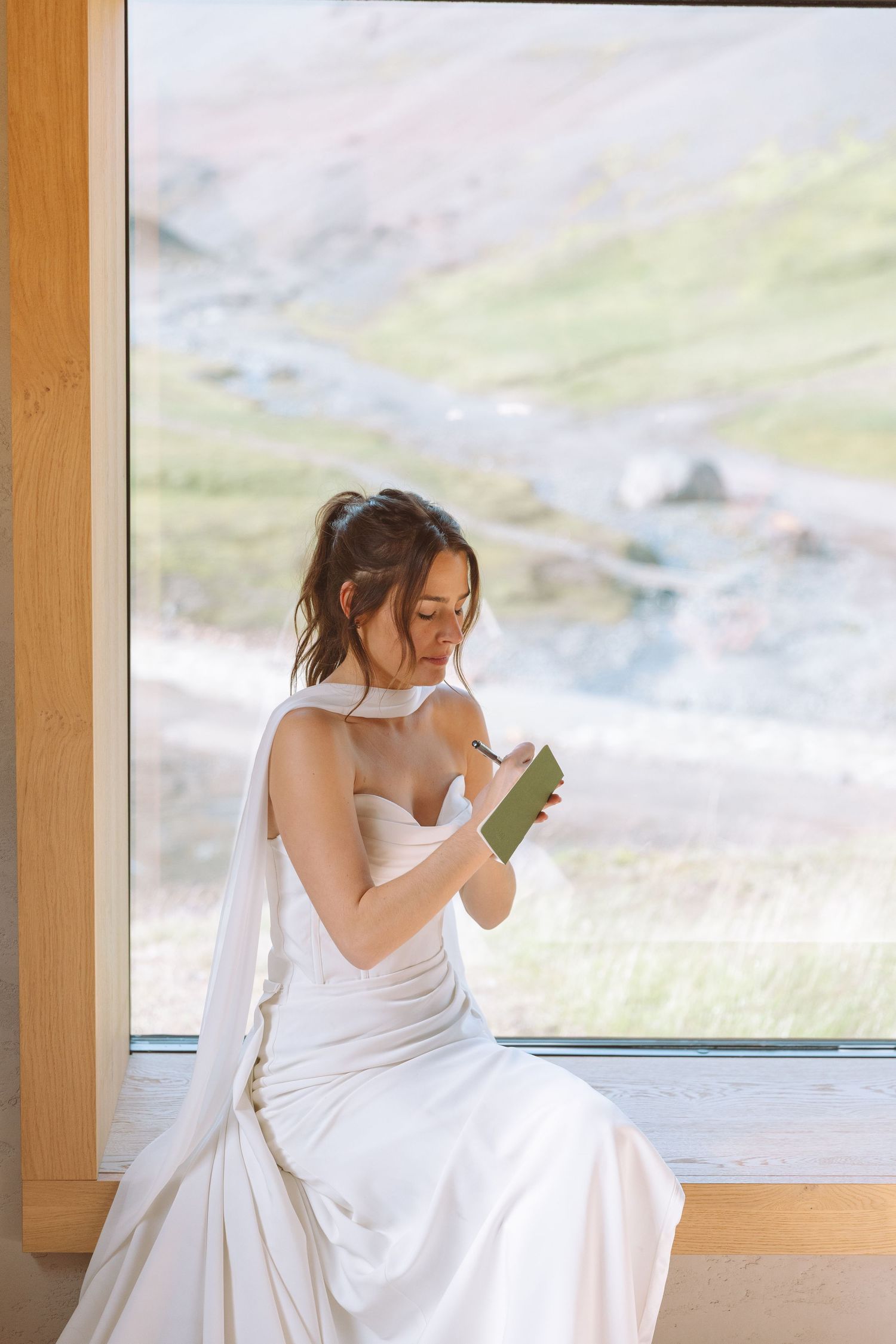A bride in a white strapless gown sits by a large window writing in a small green notebook.