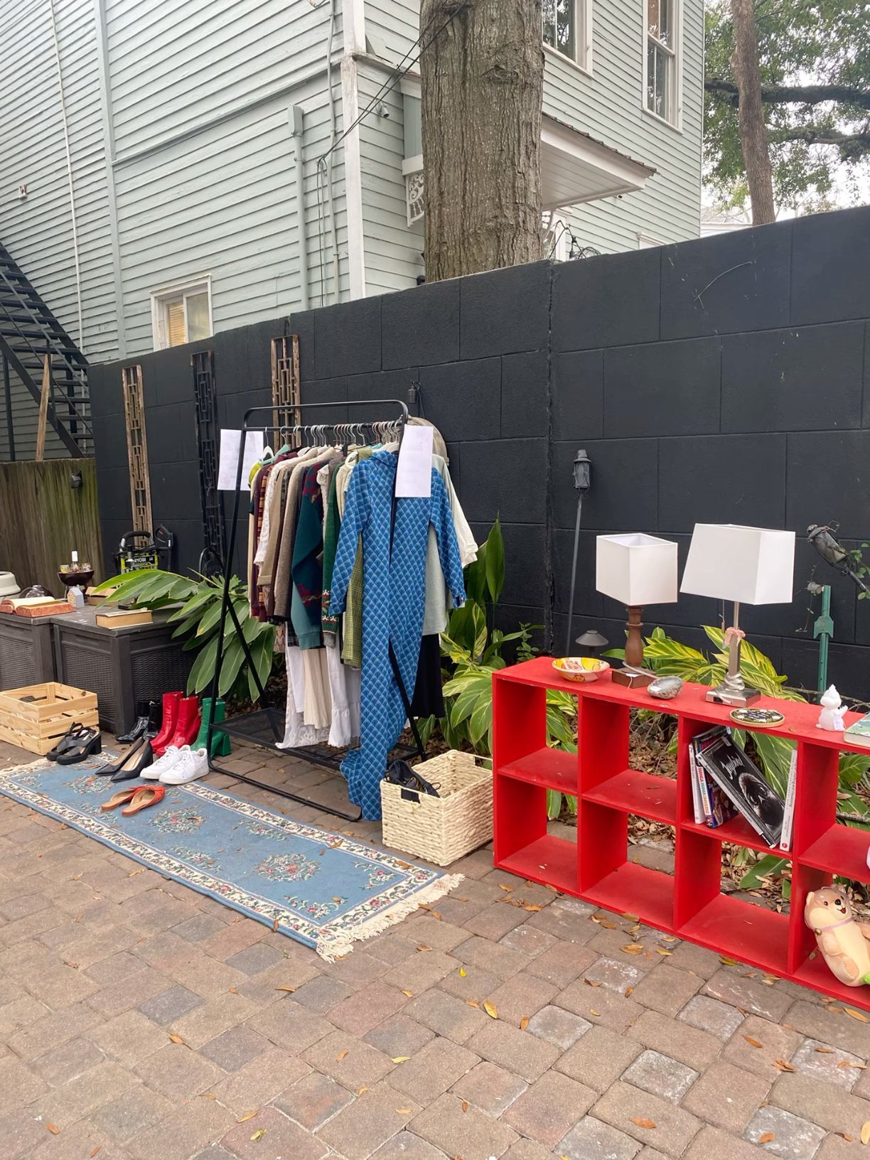 Outdoor yard sale display with clothing rack, red shelving unit, lamps, shoes, and plants against black wall.