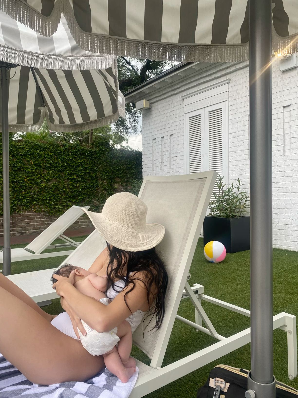 Woman in white swimsuit and sun hat relaxing on lounge chair under striped cabana in a lush backyard.