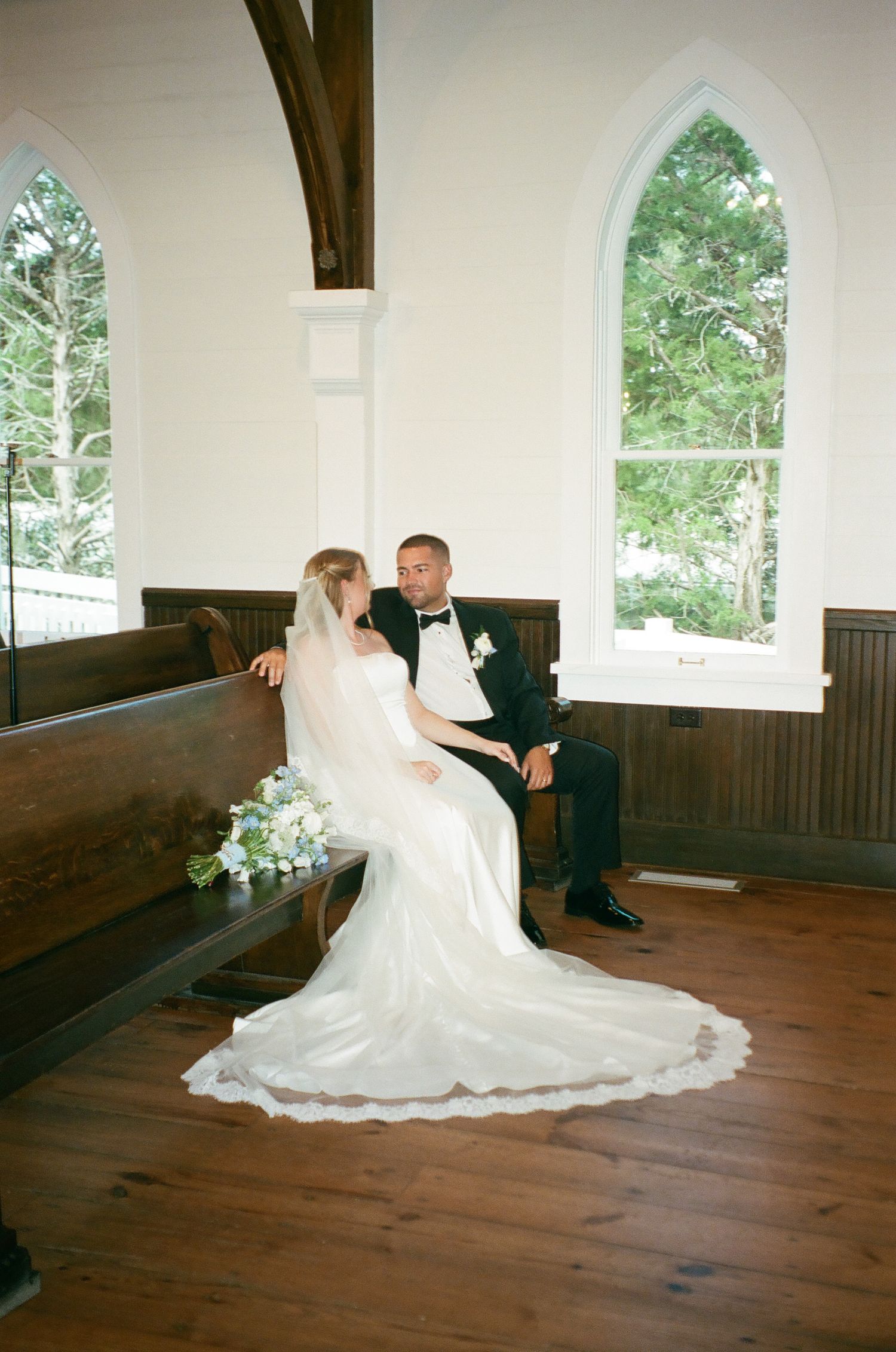 Bride in a flowing gown and groom in a tuxedo sit together on a church pew beside arched windows and a bridal bouquet.