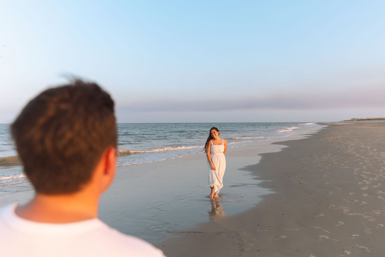 Couple on a sandy beach at sunset, woman in white dress walking toward man near ocean waves.