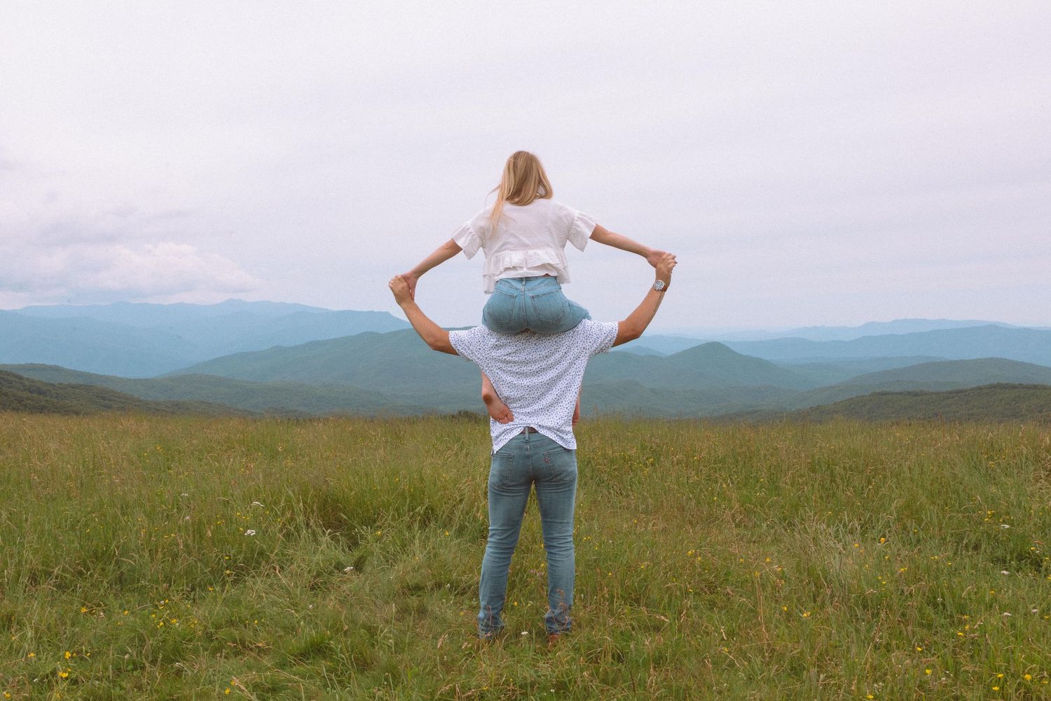 Couple standing in green mountain meadow, woman sitting on mans shoulders enjoying scenic view.