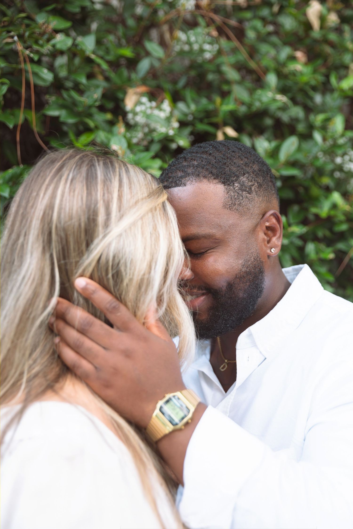 Interracial couple sharing a romantic moment, smiling and embracing outdoors with lush green foliage in the background.