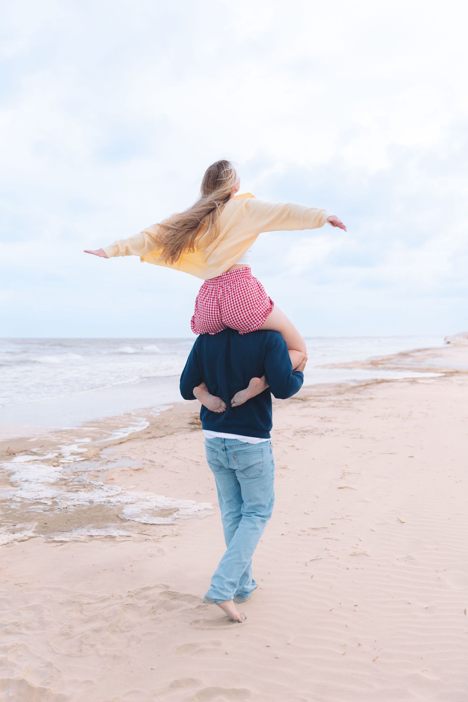 Woman sitting on mans shoulders spreading arms wide while walking on sandy beach.