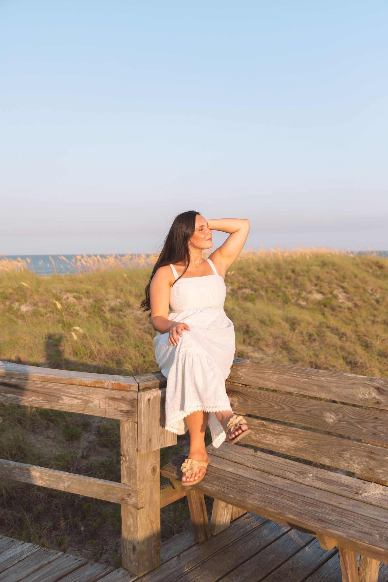 Woman in white sundress sits on wooden boardwalk railing near beach dunes at golden hour sunset.