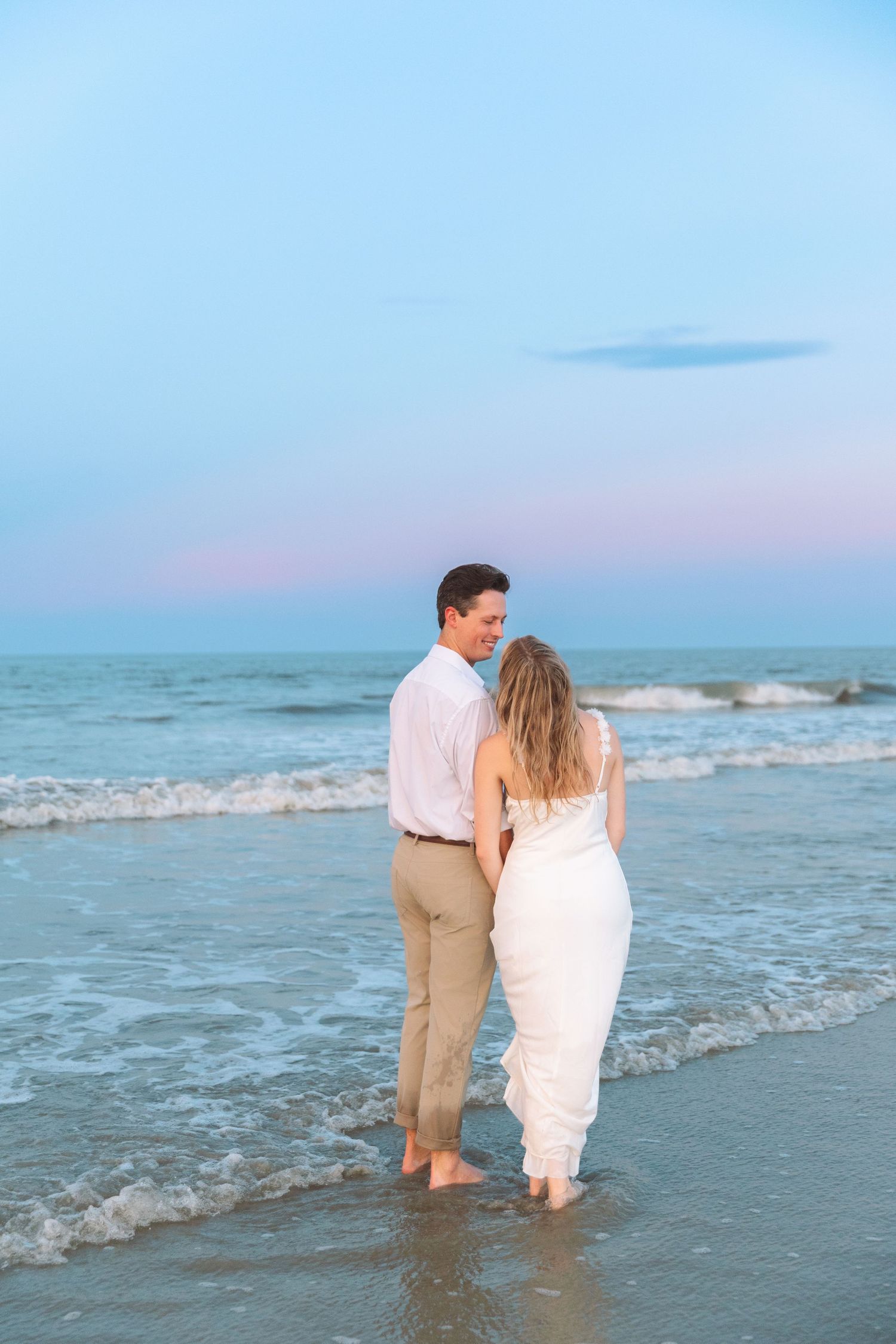 Romantic couple holding hands at the ocean's edge during a pastel sunset, woman in white dress.