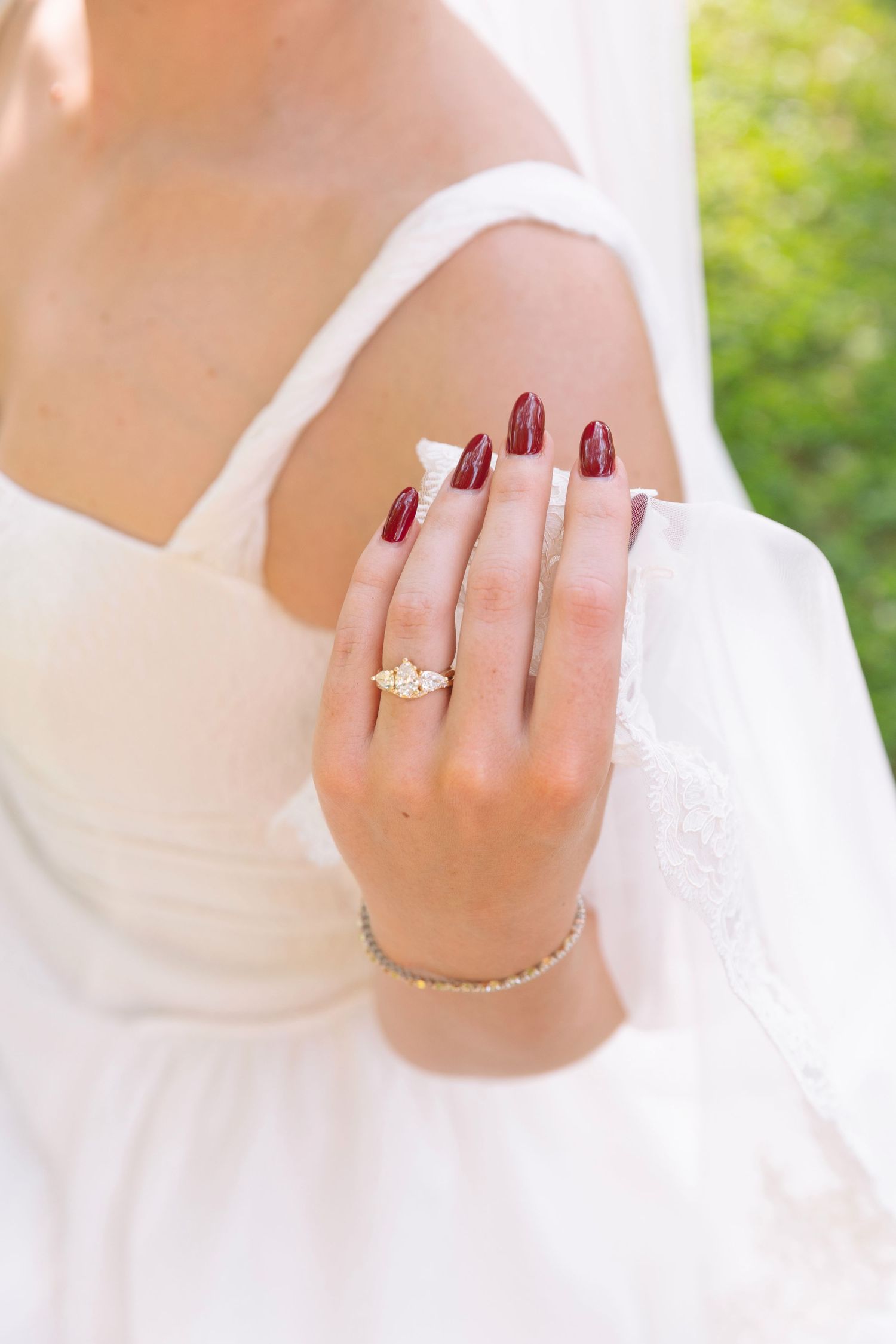 Close-up of bride's hand displaying gold engagement ring with dark red manicure against white dress.