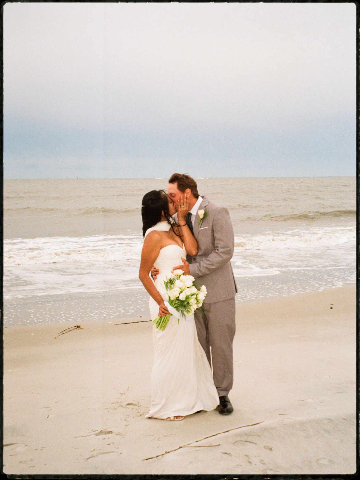 Newlyweds share romantic kiss on sandy beach holding white calla lily bouquet.