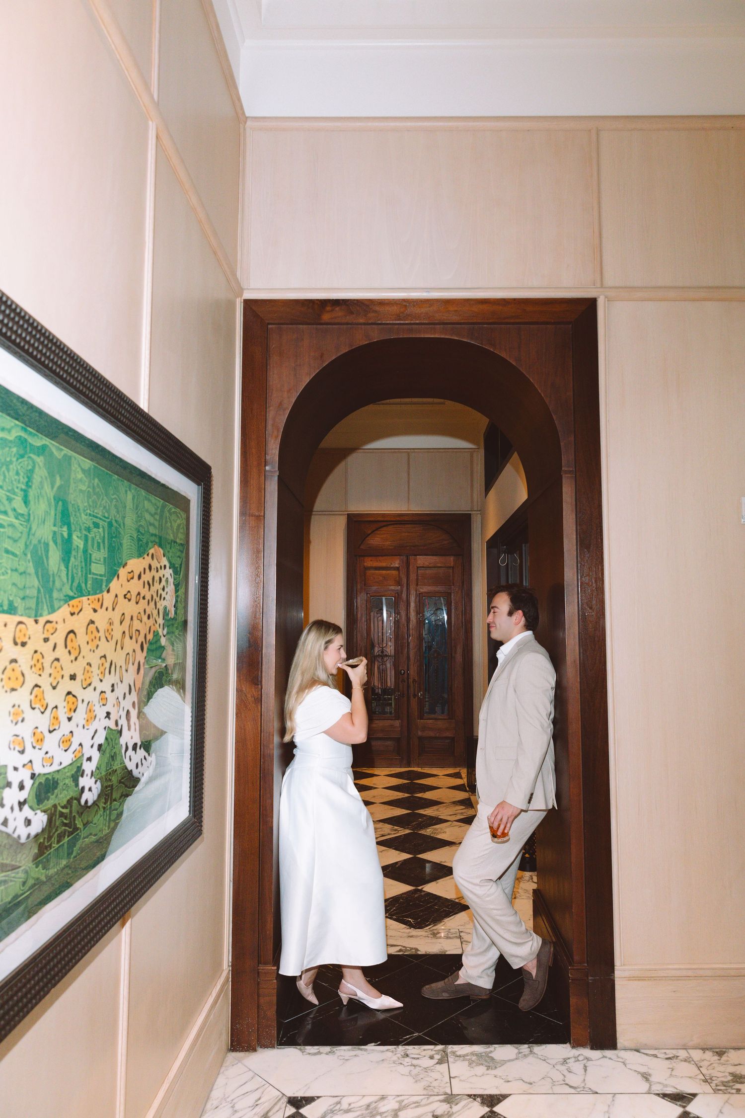 Couple in wedding attire standing under an arched doorway in an elegant foyer with checkered floors and artwork.