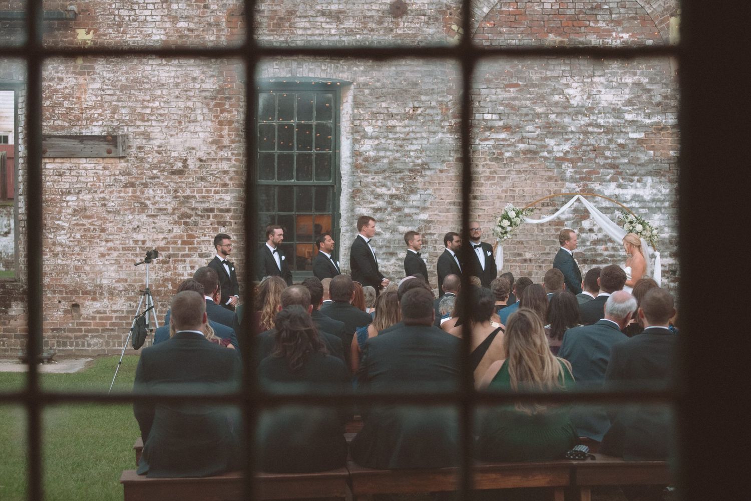 Wedding ceremony viewed through a window at a rustic brick venue with seated guests.