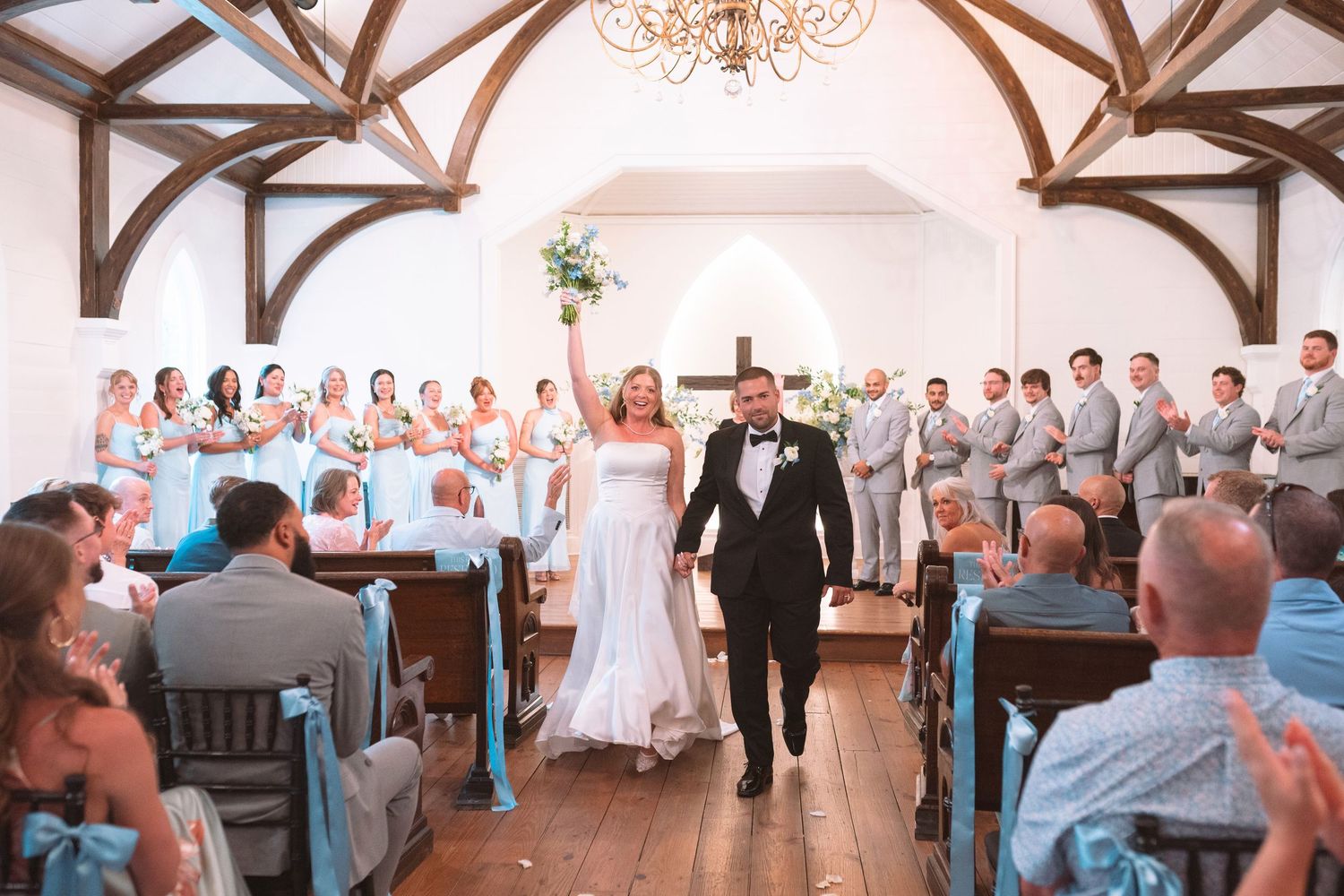Newlyweds celebrating as they walk down the aisle in a white chapel surrounded by bridesmaids and groomsmen.