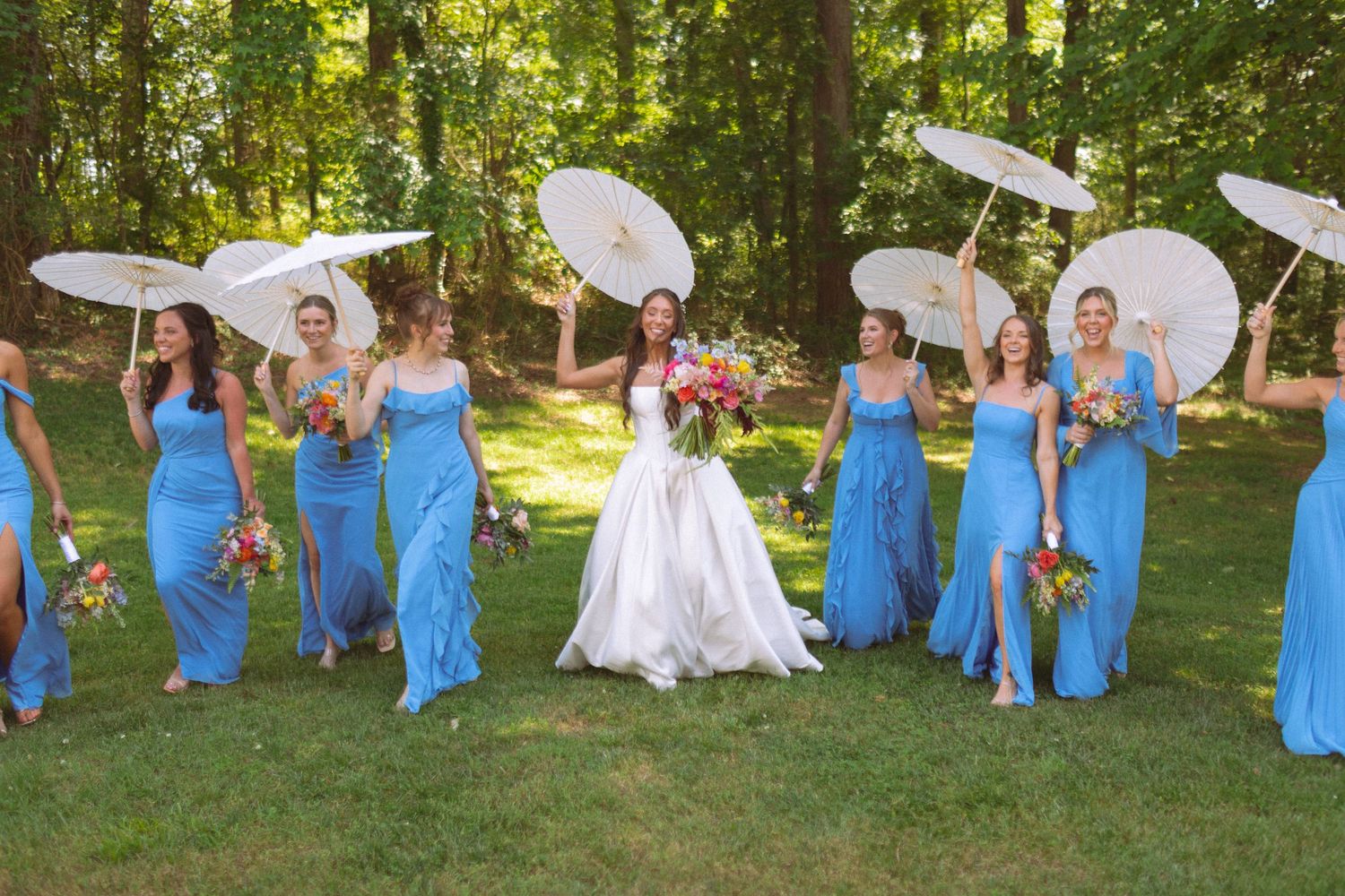 Bridesmaids in blue dresses holding white parasols walk with bride outdoors at garden wedding.