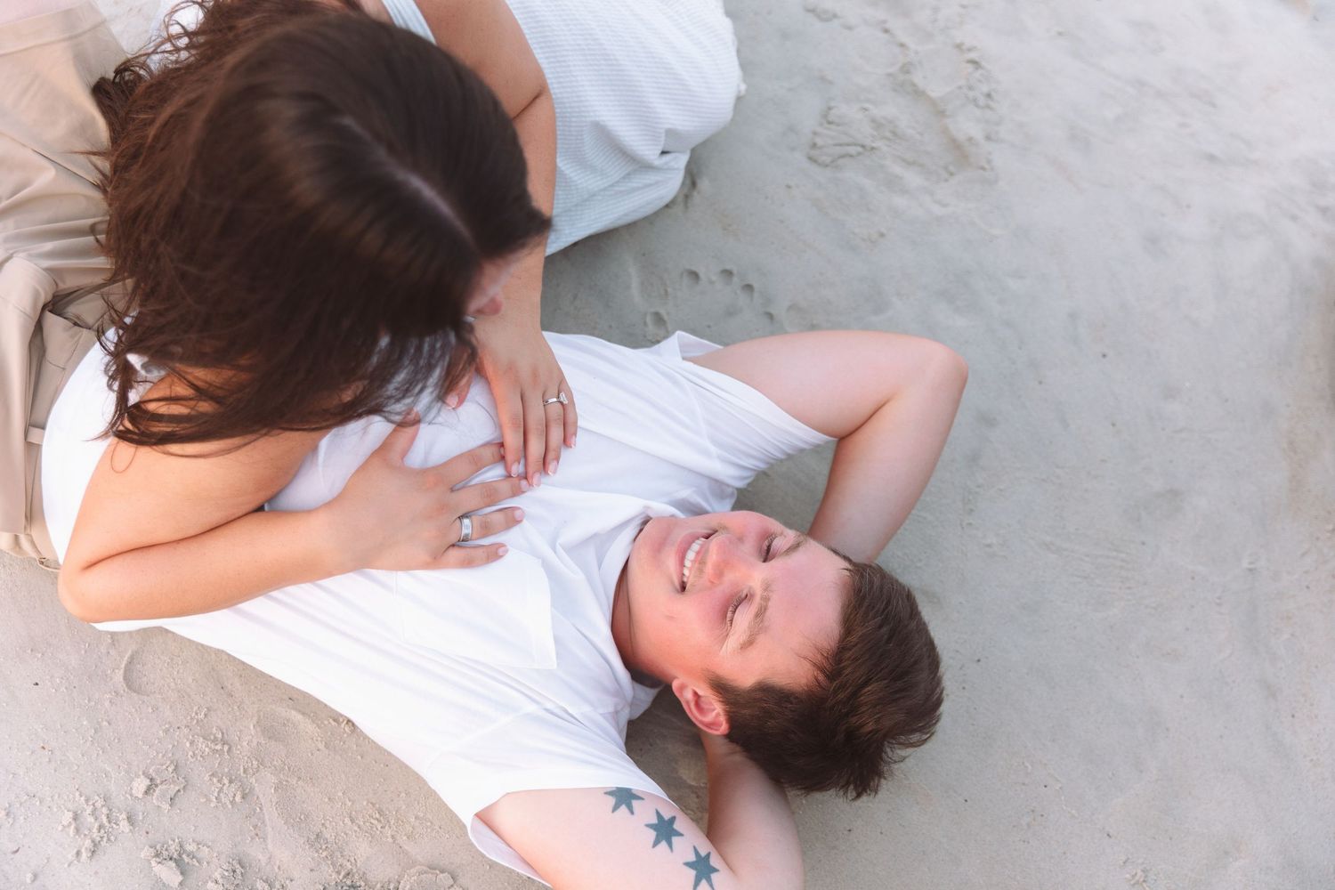 Couple relaxing together on sandy beach, man lying down smiling with tattoo, woman leaning over him.