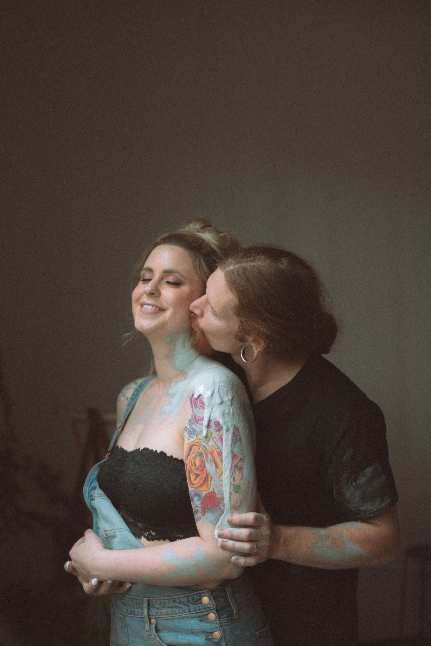 Woman with colorful tattoos smiling as partner embraces and kisses her cheek indoors.