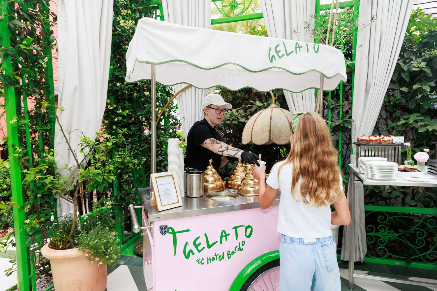 A man serves gelato from a charming pink and white outdoor cart surrounded by lush greenery to a young girl.