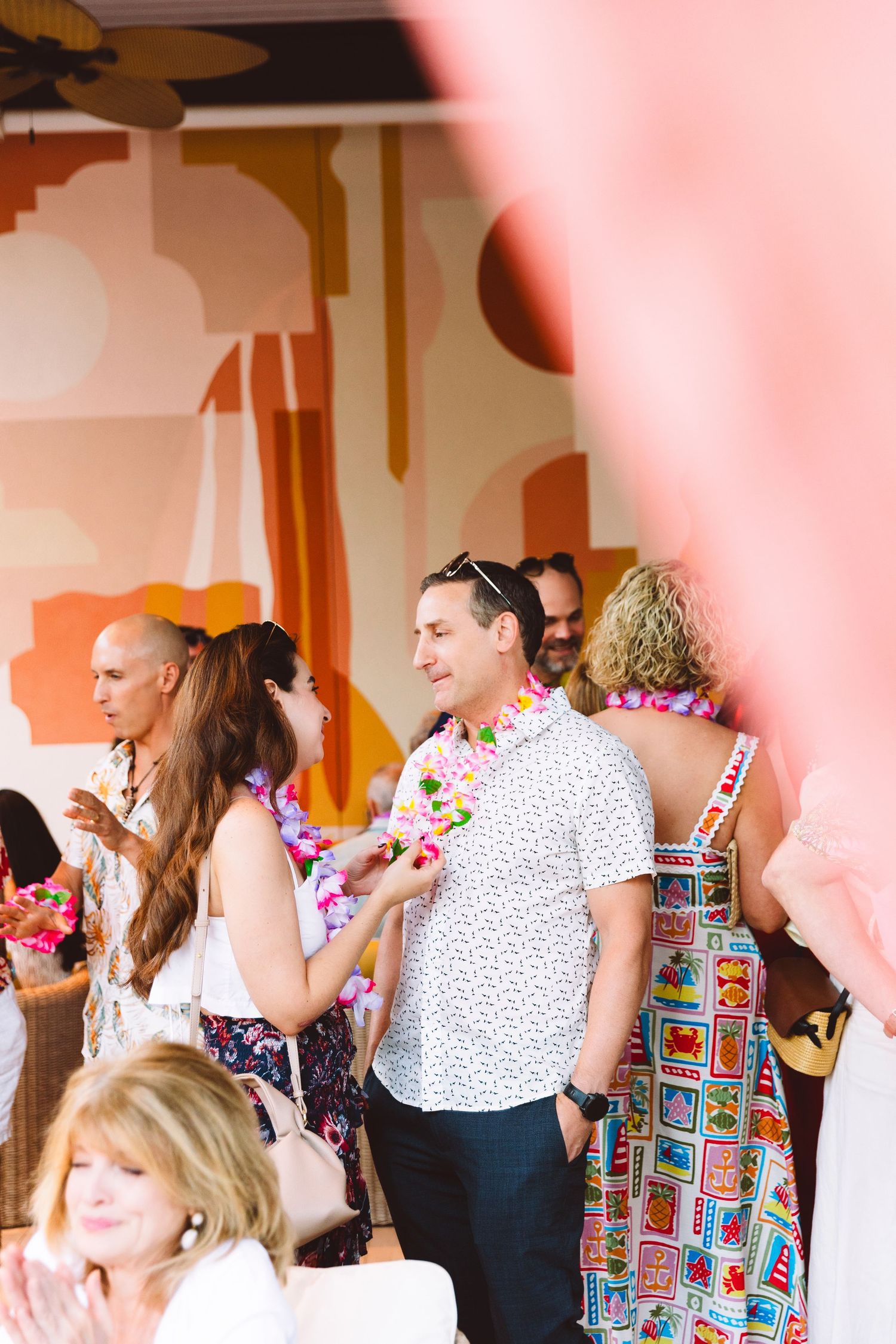 Guests wearing colorful leis mingle at a tropical-themed party with vibrant pink and orange decor.