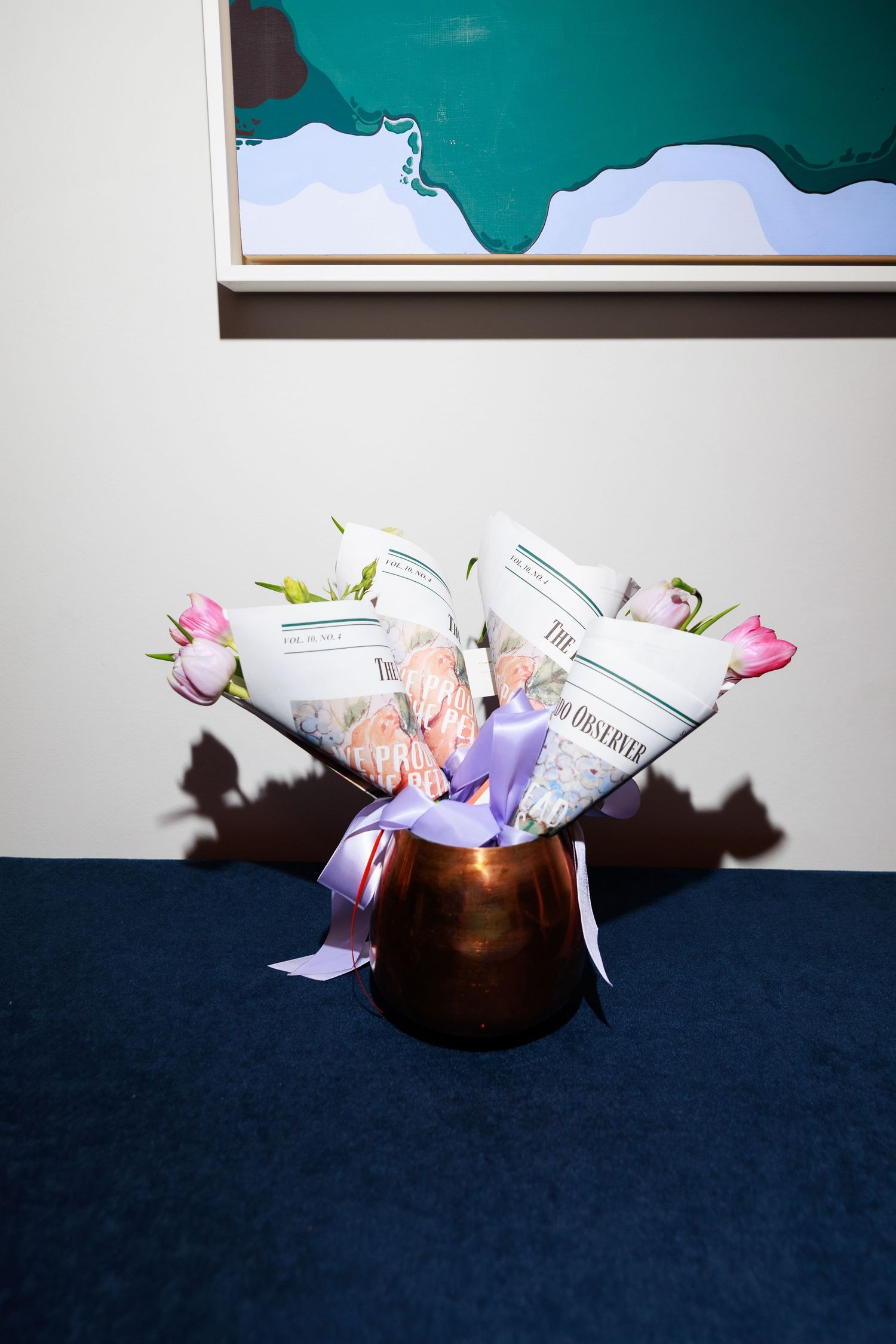 Copper vase with pink tulips wrapped in newspaper, tied with lavender ribbon, on blue surface near abstract wall art.