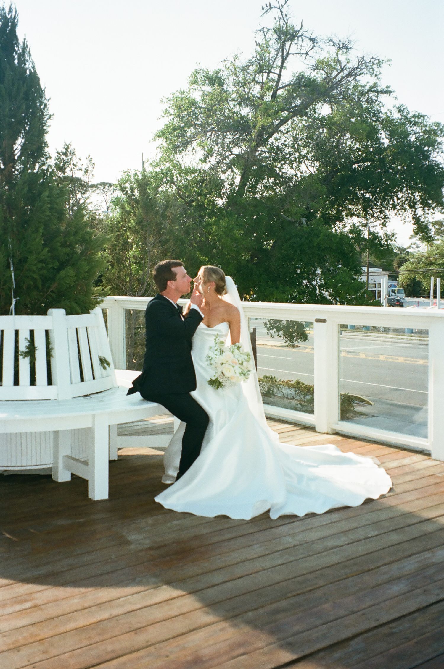 Bride and groom share a romantic kiss on a wooden deck with white bench and lush trees at their outdoor wedding.