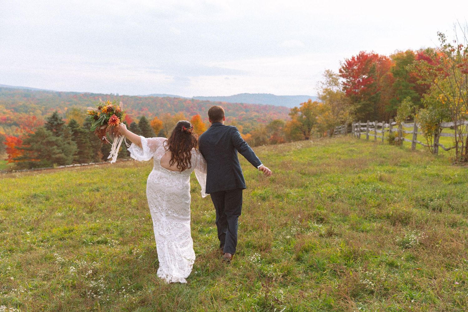 Bride and groom walking hand in hand across green meadow with stunning fall foliage landscape.