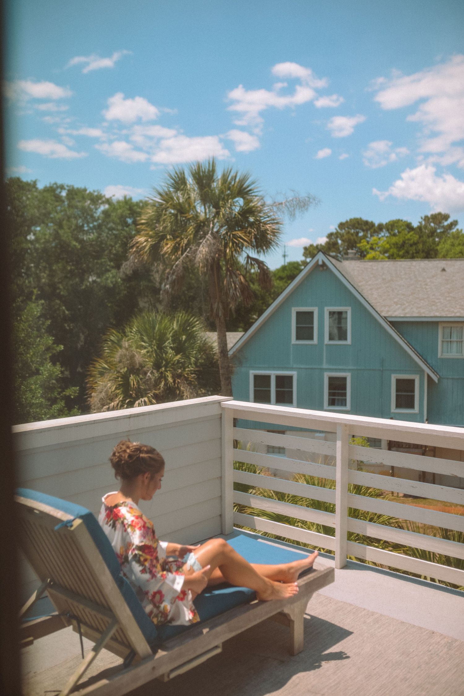 Woman in floral robe sitting on lounge chair on sunny deck with palm trees and teal house backdrop.
