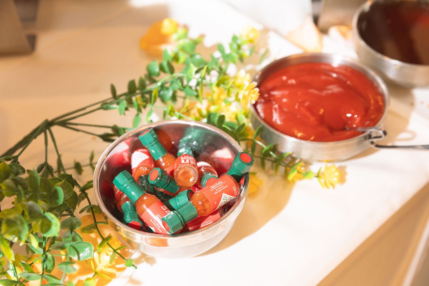 Bowl of colorful wrapped candies beside a bowl of red sauce and yellow flowers on a white table.