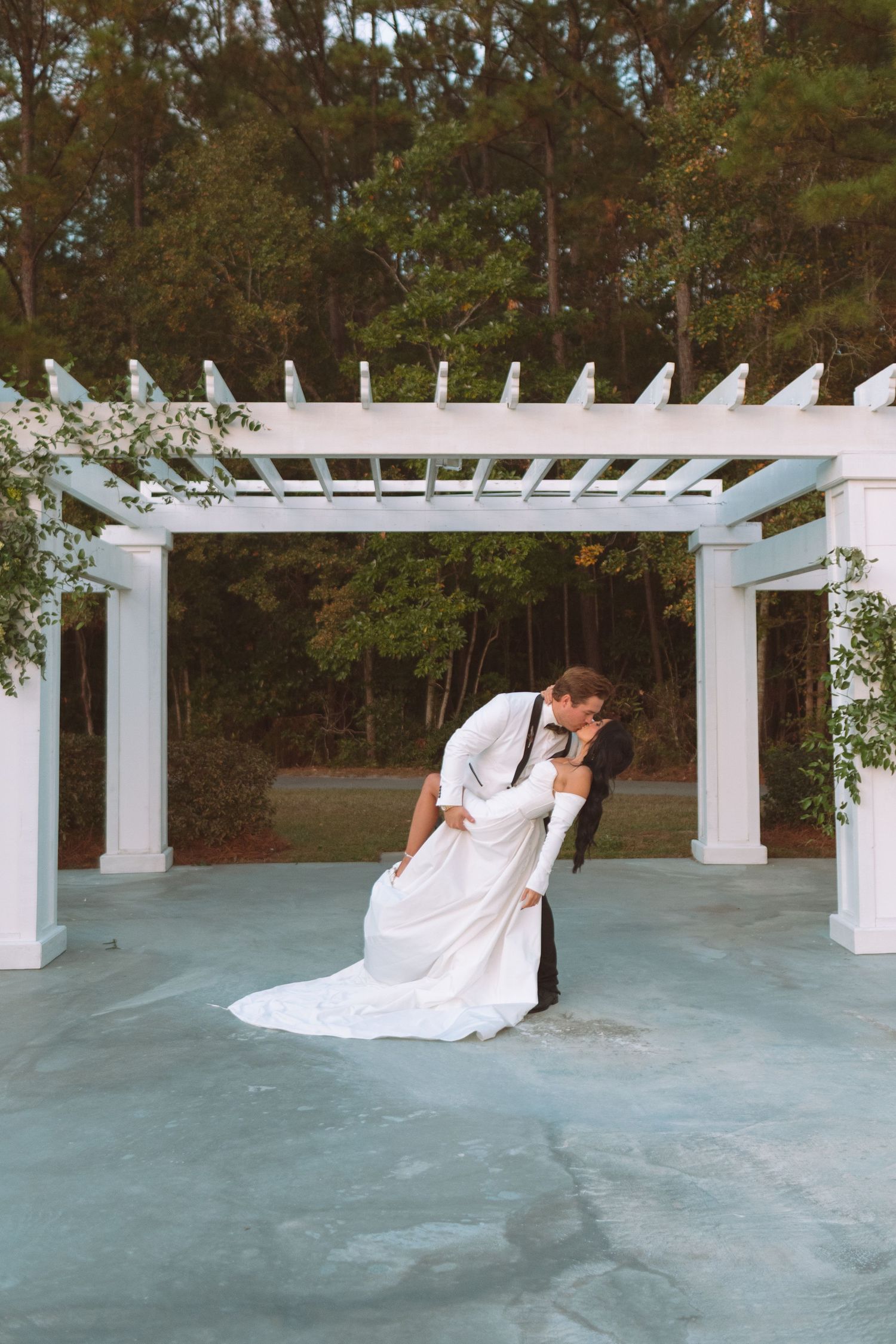Bride and groom share a romantic dip kiss under a white pergola surrounded by lush trees.