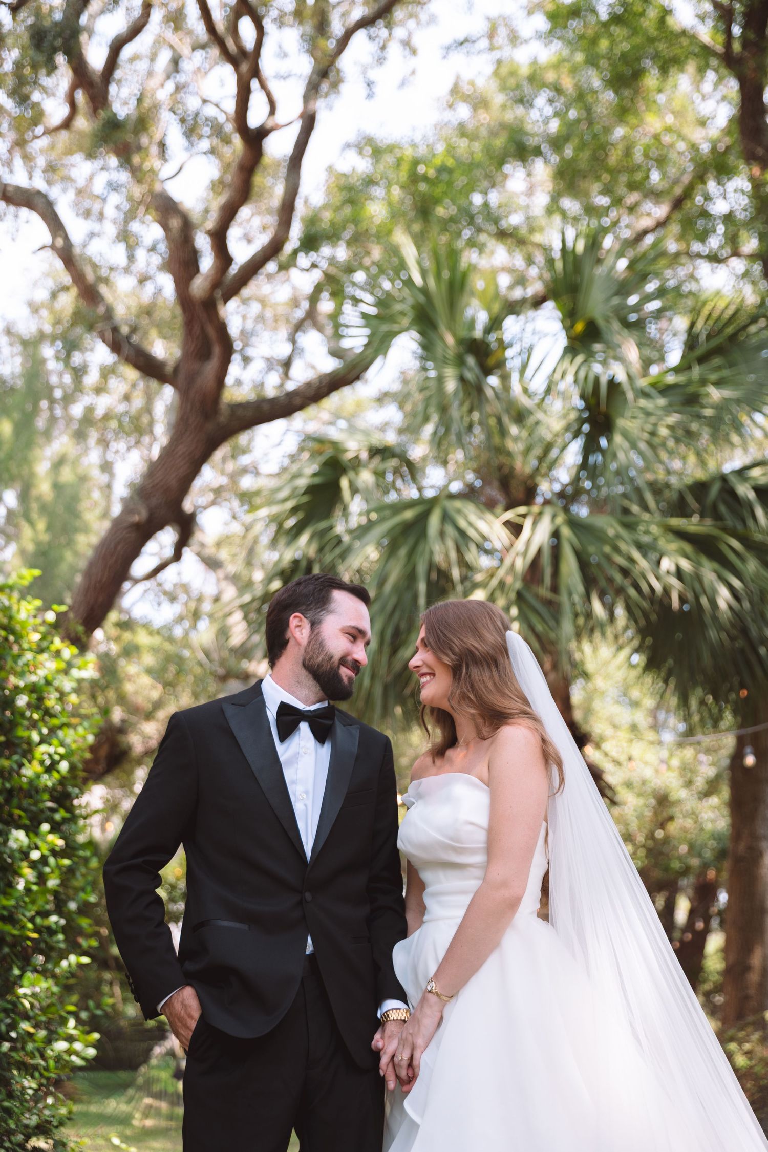 Bride and groom smiling at each other in a lush garden setting, groom in black tuxedo, bride in strapless gown with veil.