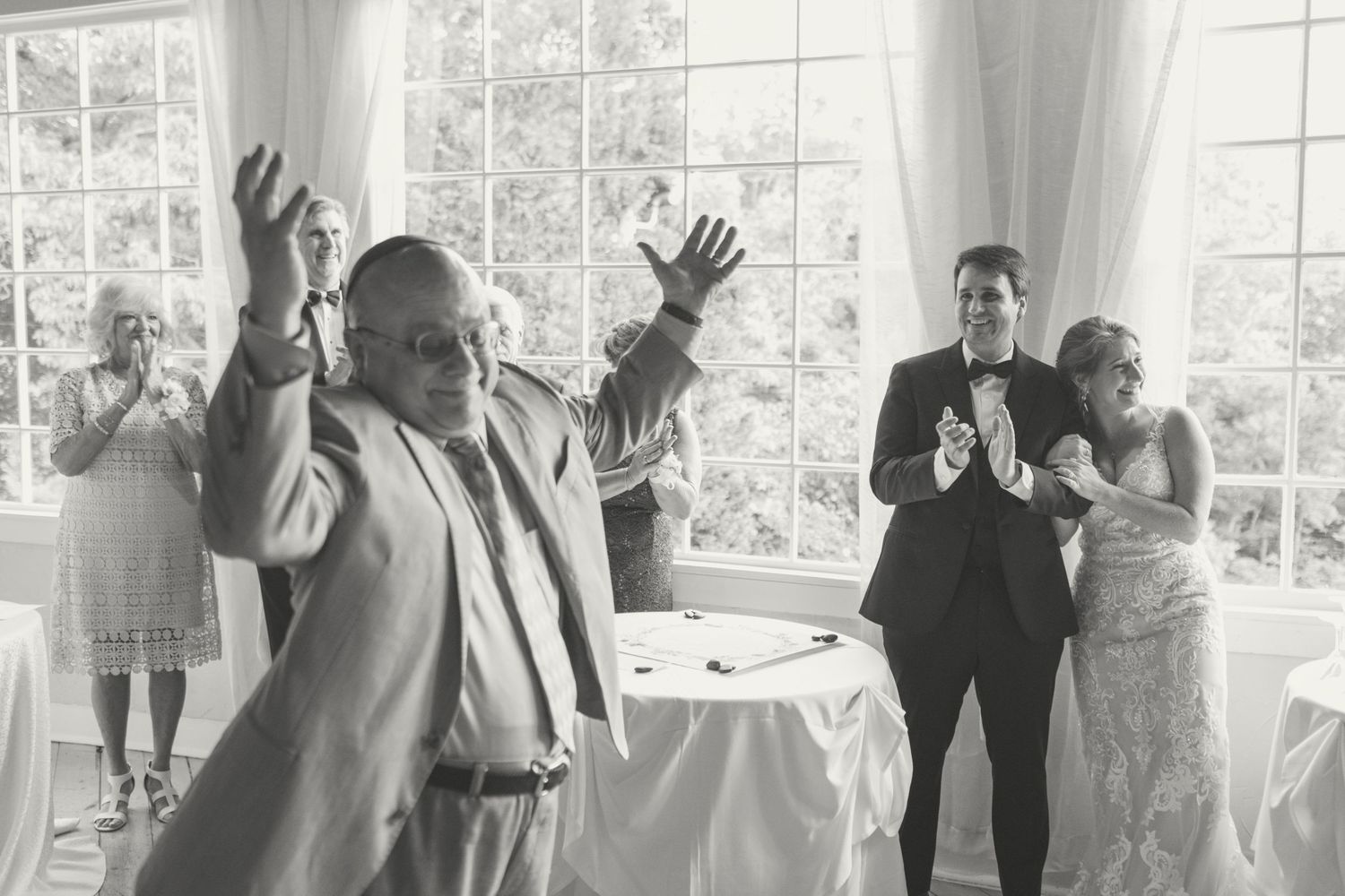 Black and white photo of wedding guests dancing and celebrating at an indoor wedding reception.