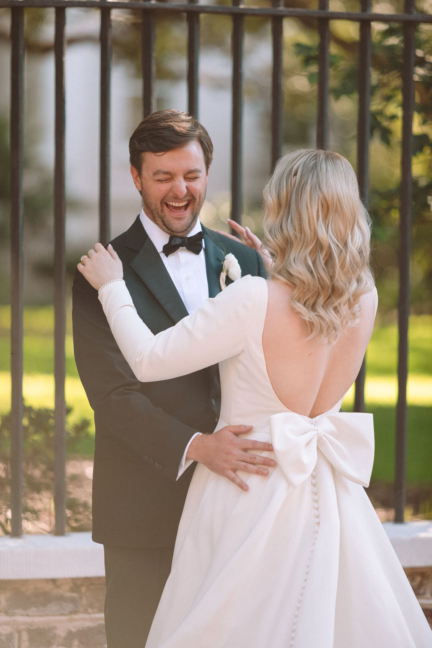 Joyful groom in black tuxedo laughing as bride in backless bow wedding dress embraces him outdoors.