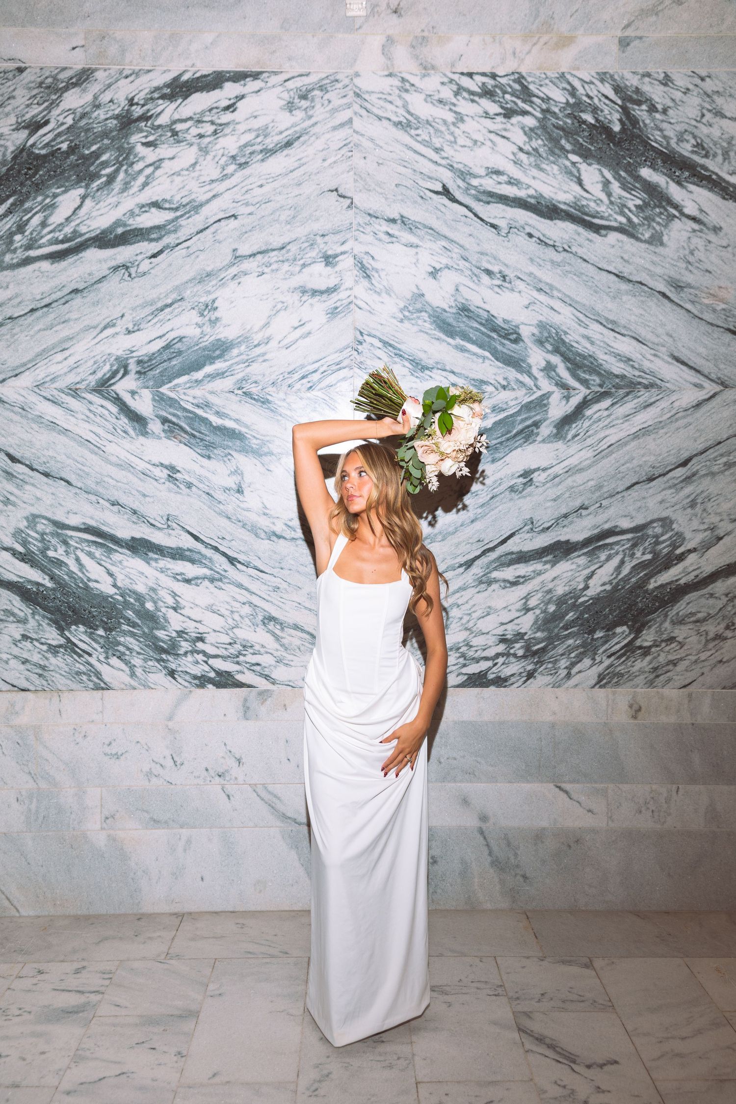 Bride in white gown holds floral bouquet overhead before marble wall.