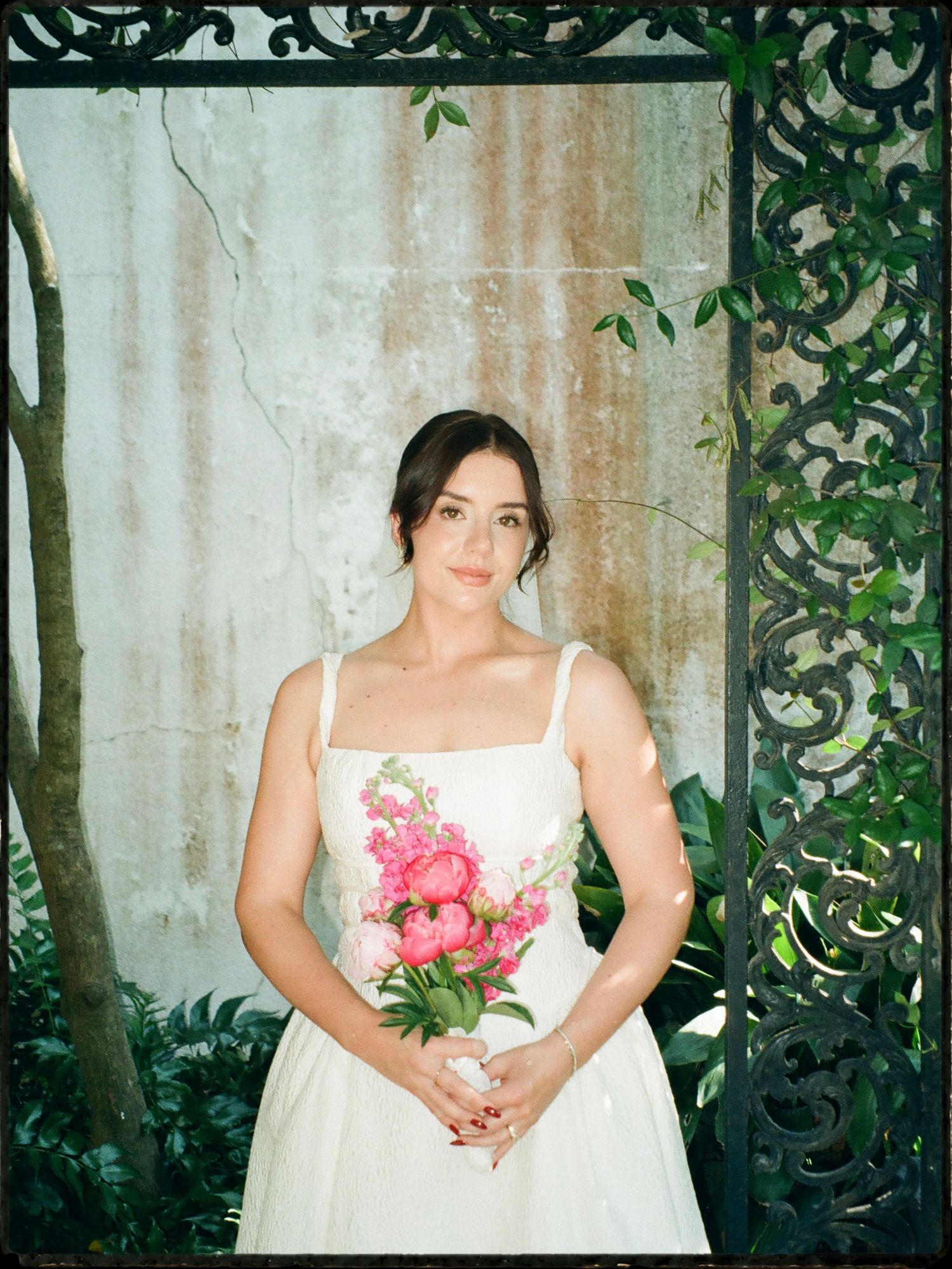 Bride in white floral wedding dress holding pink bouquet framed by ornate black iron gate.