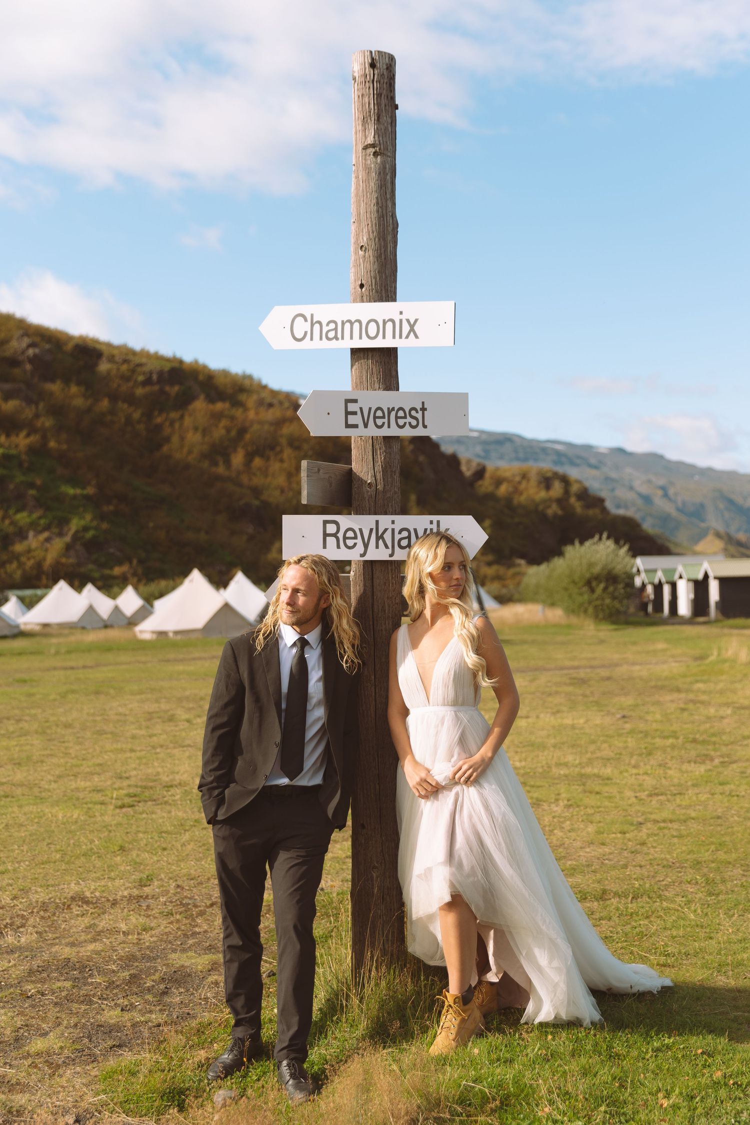 Bride and groom stand by a rustic wooden signpost pointing to Chamonix, Everest, and Reykjavik at an outdoor wedding.