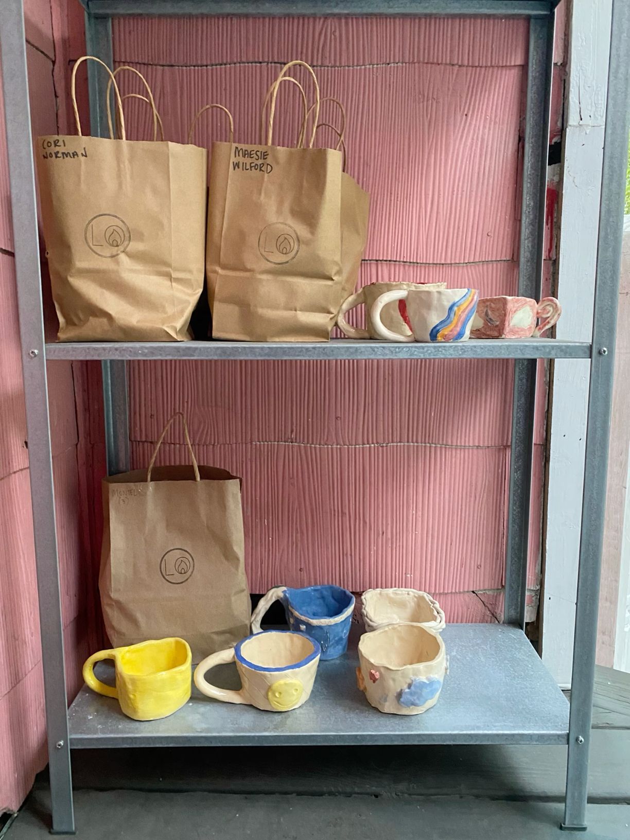 Metal shelf displaying handmade ceramic mugs in yellow, blue, and multicolor beside brown paper bags.