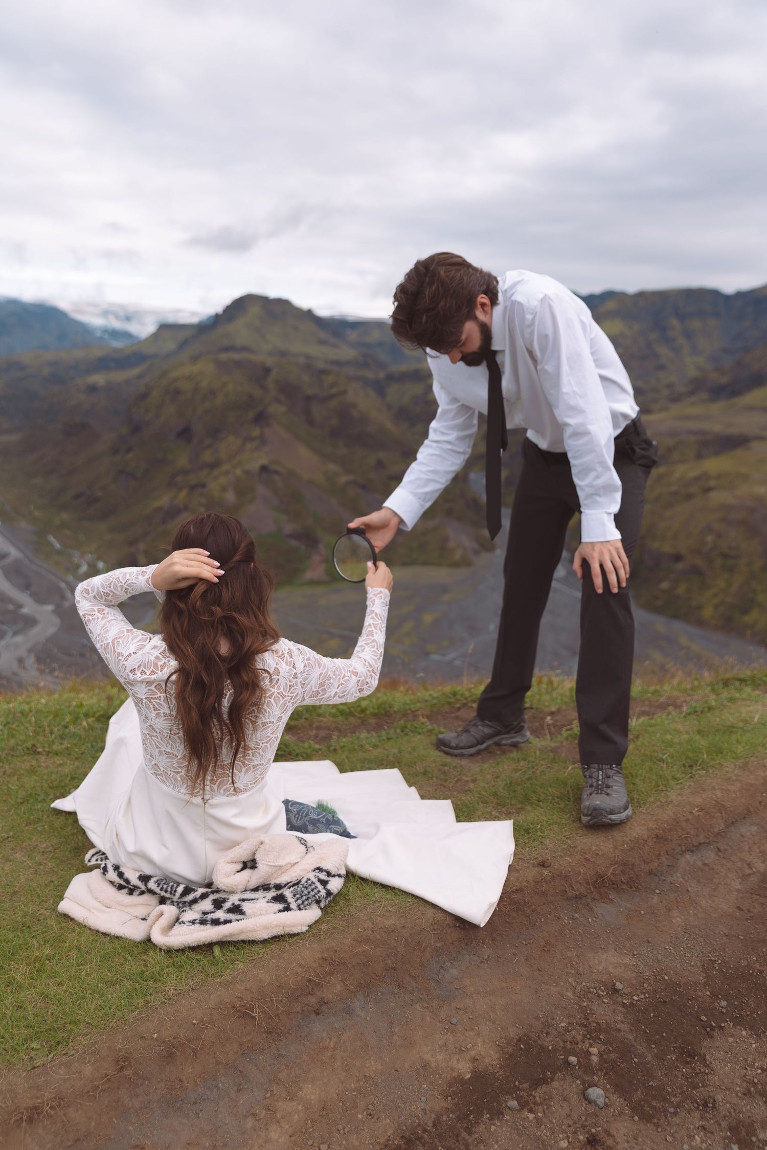 Groom holding mirror for bride sitting on blanket atop scenic Icelandic mountain ridge.
